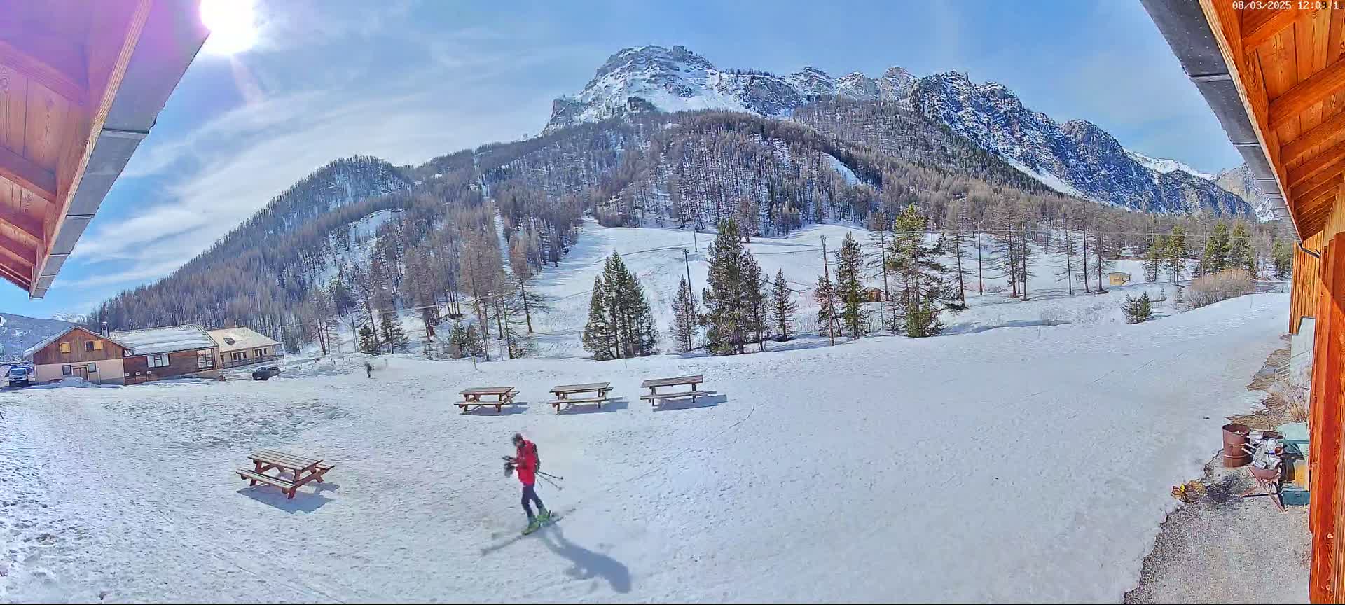 A person skis across a snow-covered area with picnic tables and buildings in front of a mountain range under a sunny sky.