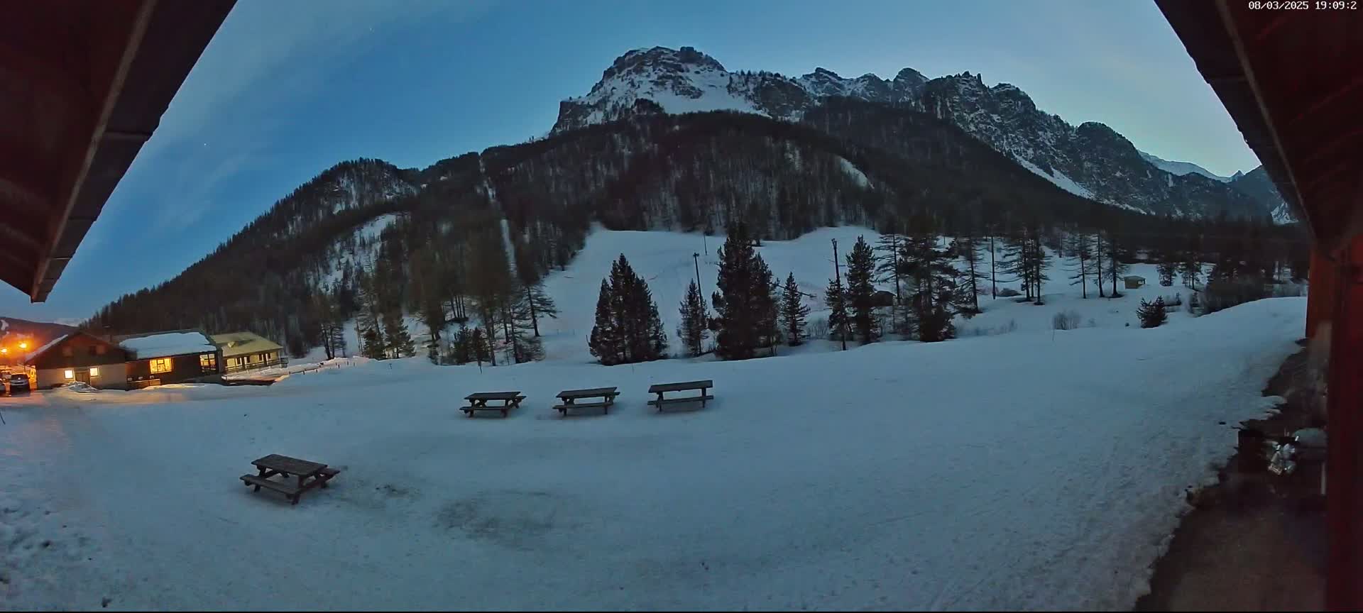 A snow-covered landscape at twilight features several picnic tables in the foreground, a small building in the middle ground, and a large mountain in the background.