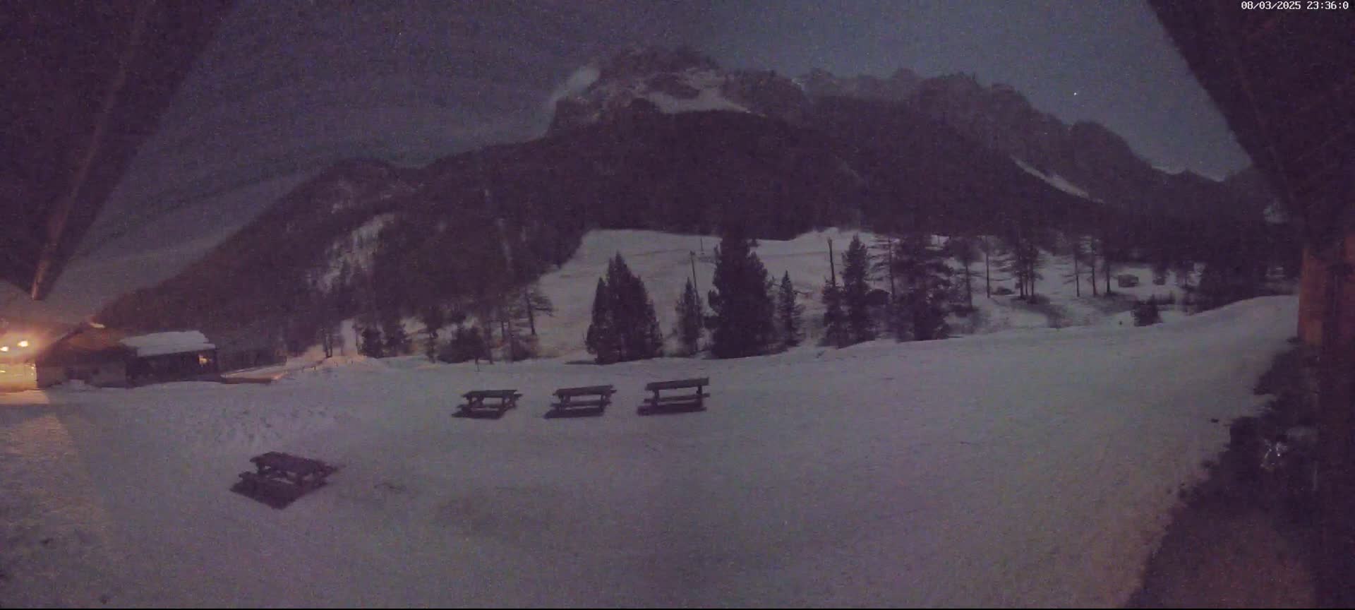 A snow-covered landscape at night shows a line of picnic tables in the foreground, a small building to the left, and a mountain range in the background under a dark, possibly snowy sky.
