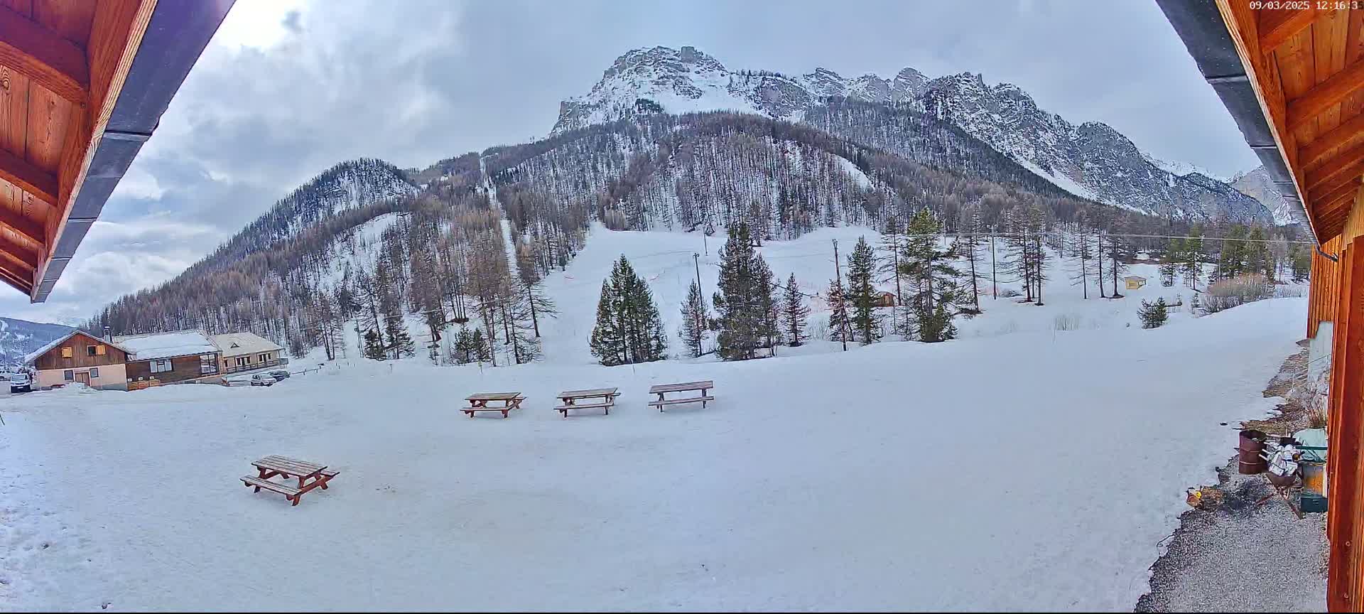 A snowy landscape with several picnic tables sits in front of a snow-covered mountain, under a cloudy sky.