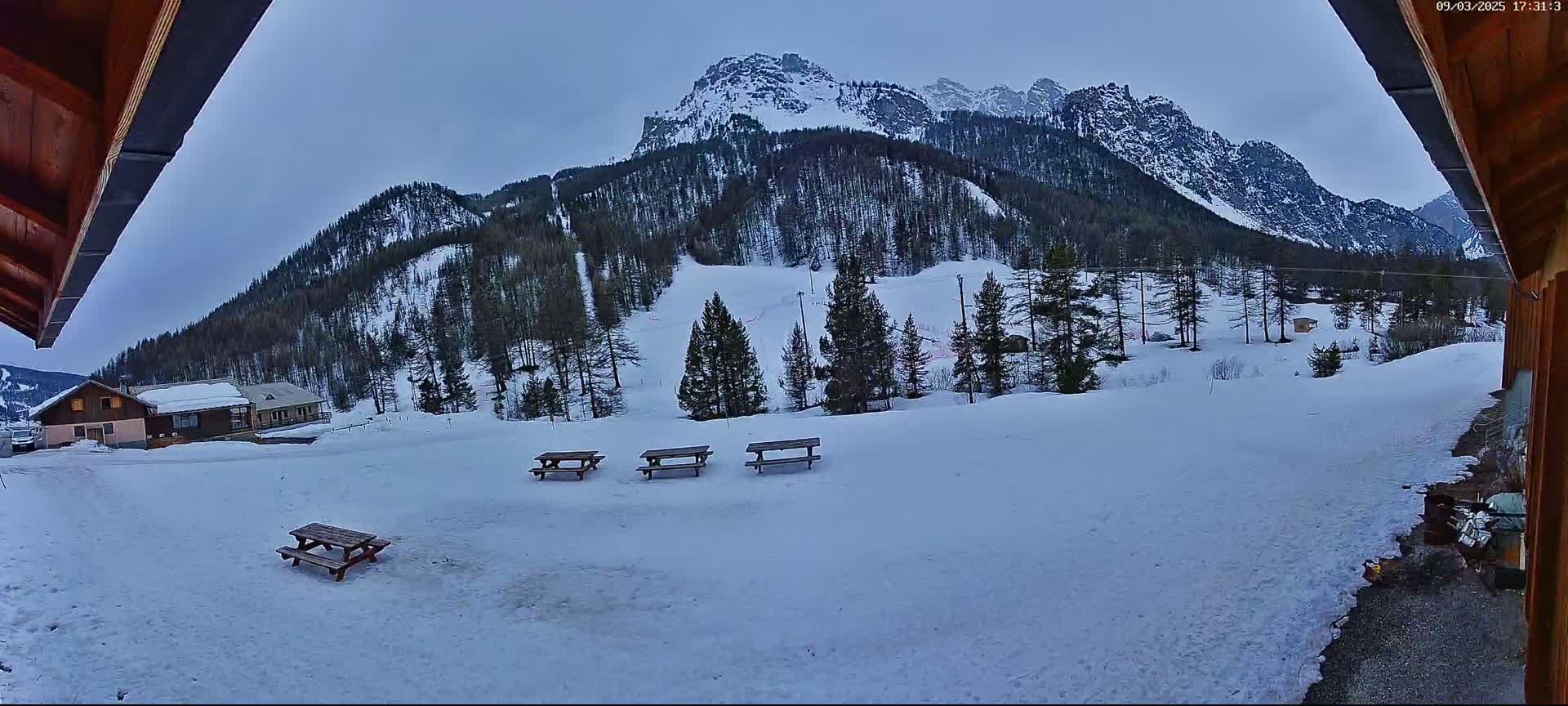 A snowy landscape with several picnic tables in the foreground and a snow-covered mountain in the background under a cloudy sky.