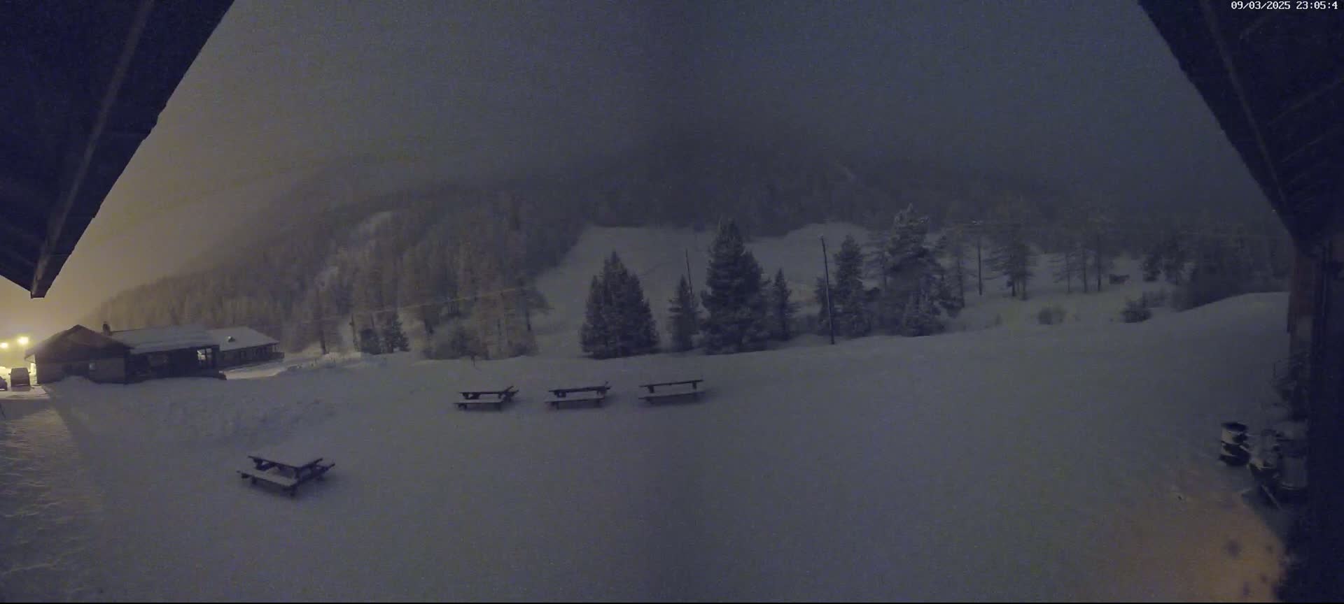 A snow-covered landscape at night, with a few picnic tables and a building visible under a dark, hazy sky.