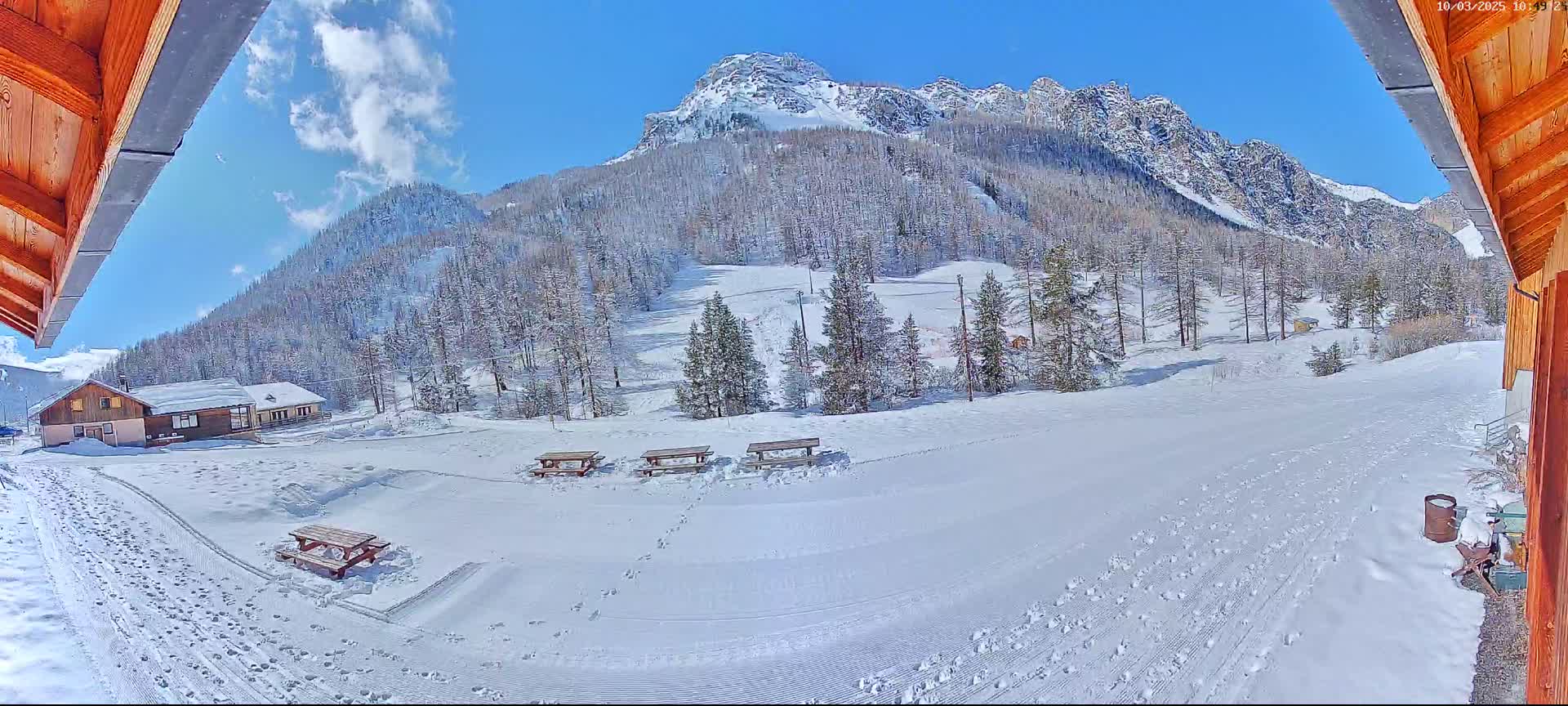A snow-covered landscape with several picnic tables, a building, and a mountain in the background under a mostly sunny sky.