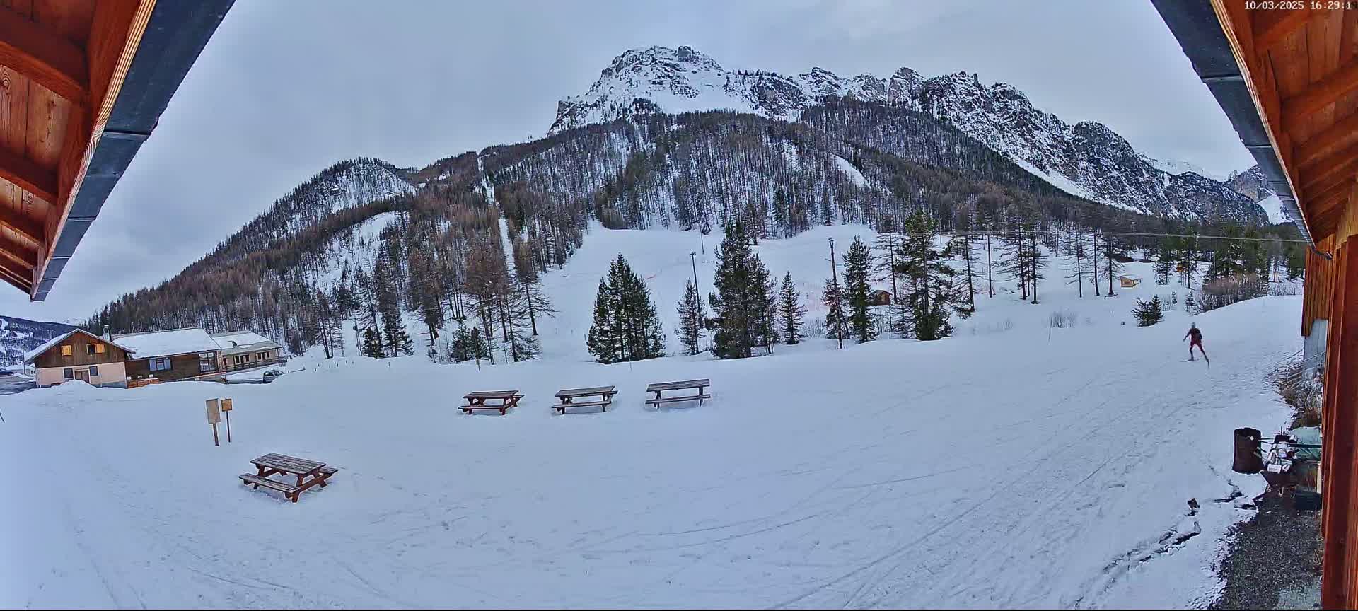 A snowy, overcast scene shows a person cross-country skiing in front of a snow-covered mountain with several picnic tables and a building in the foreground.