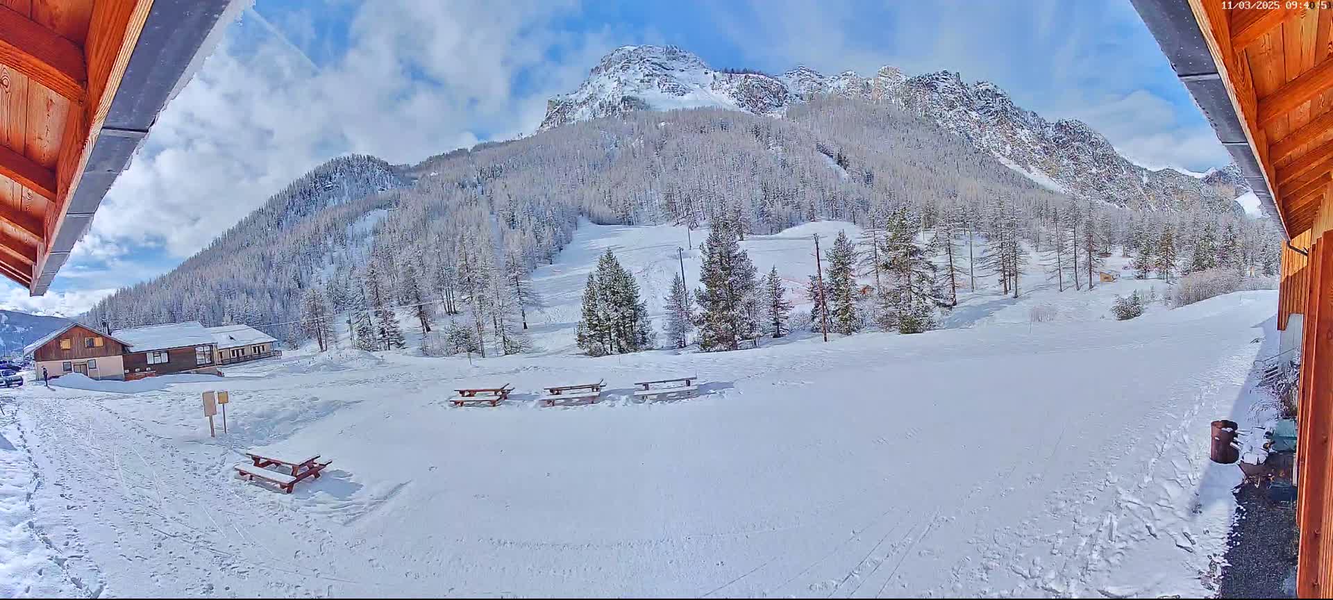 A snow-covered mountainside with several picnic tables and a small building is visible from under a wooden overhang on a partly cloudy day.