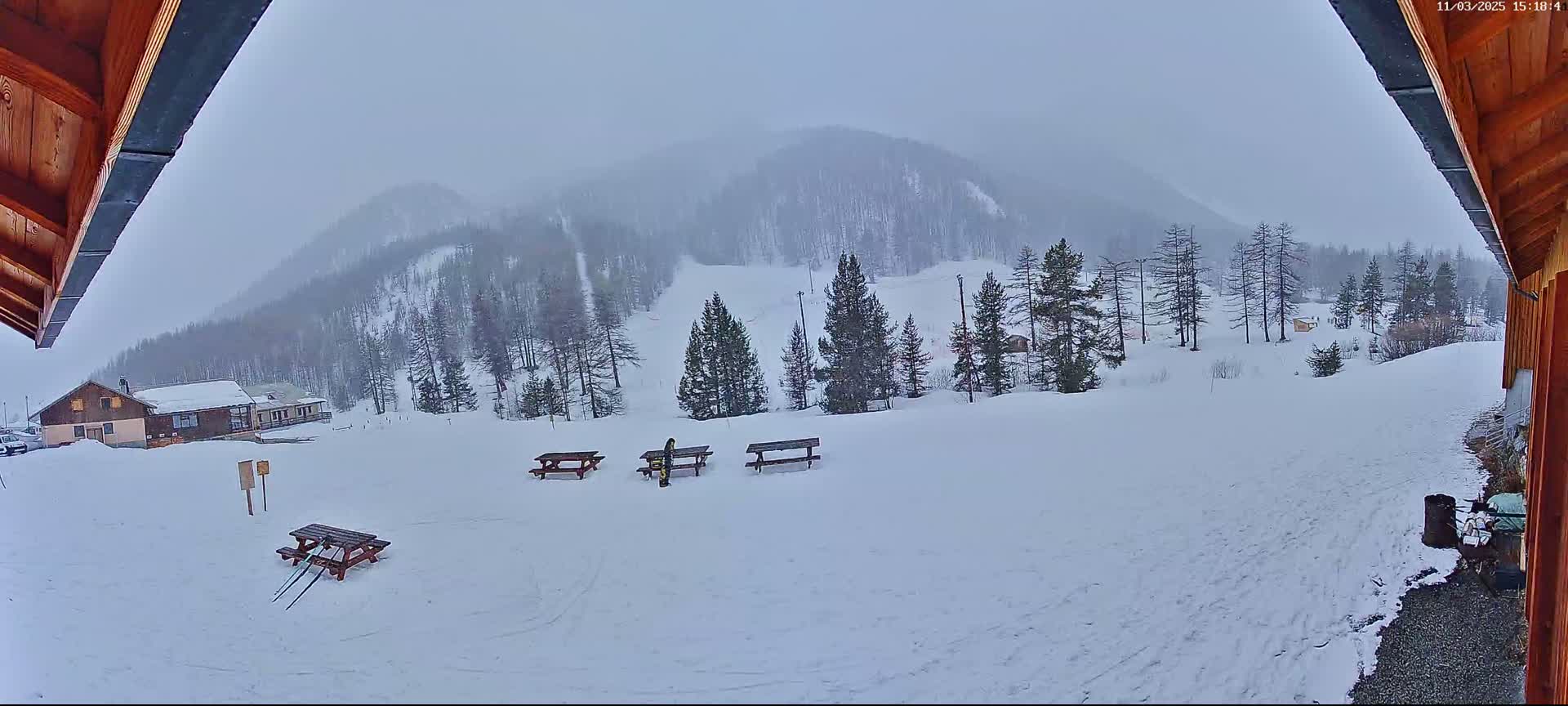 A snowy landscape with several picnic tables, a distant building, and a few evergreen trees under an overcast sky.