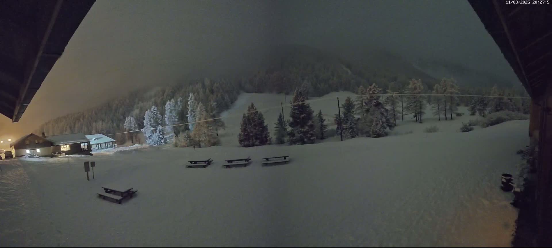 A snow-covered landscape at night, featuring a building, several picnic tables, and a forest on a hill under a dark, overcast sky.