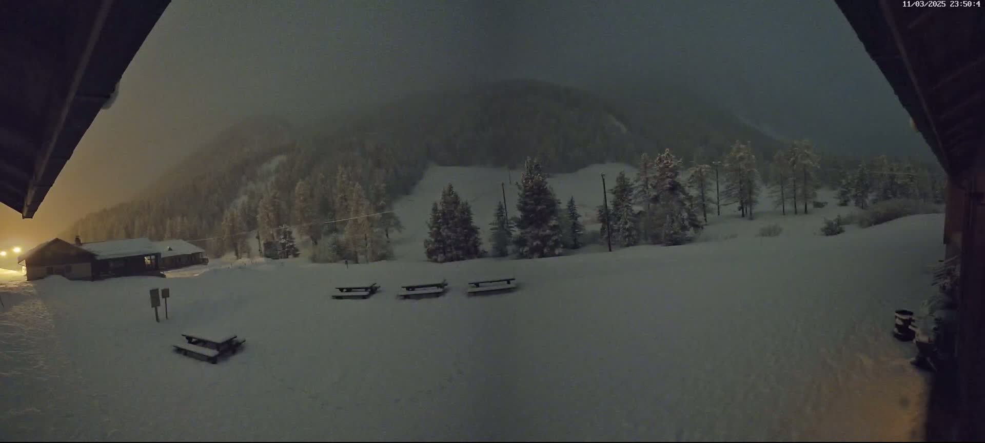 A snow-covered landscape at night, featuring several picnic tables and a small building in the foreground, with a snow-covered hillside and dark forest in the background under a hazy sky.