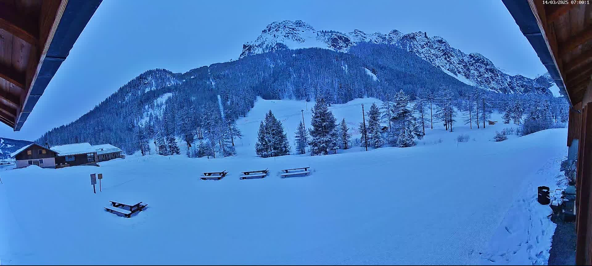 A snow-covered landscape with several picnic tables, a few buildings, and a mountain in the background under a twilight sky.