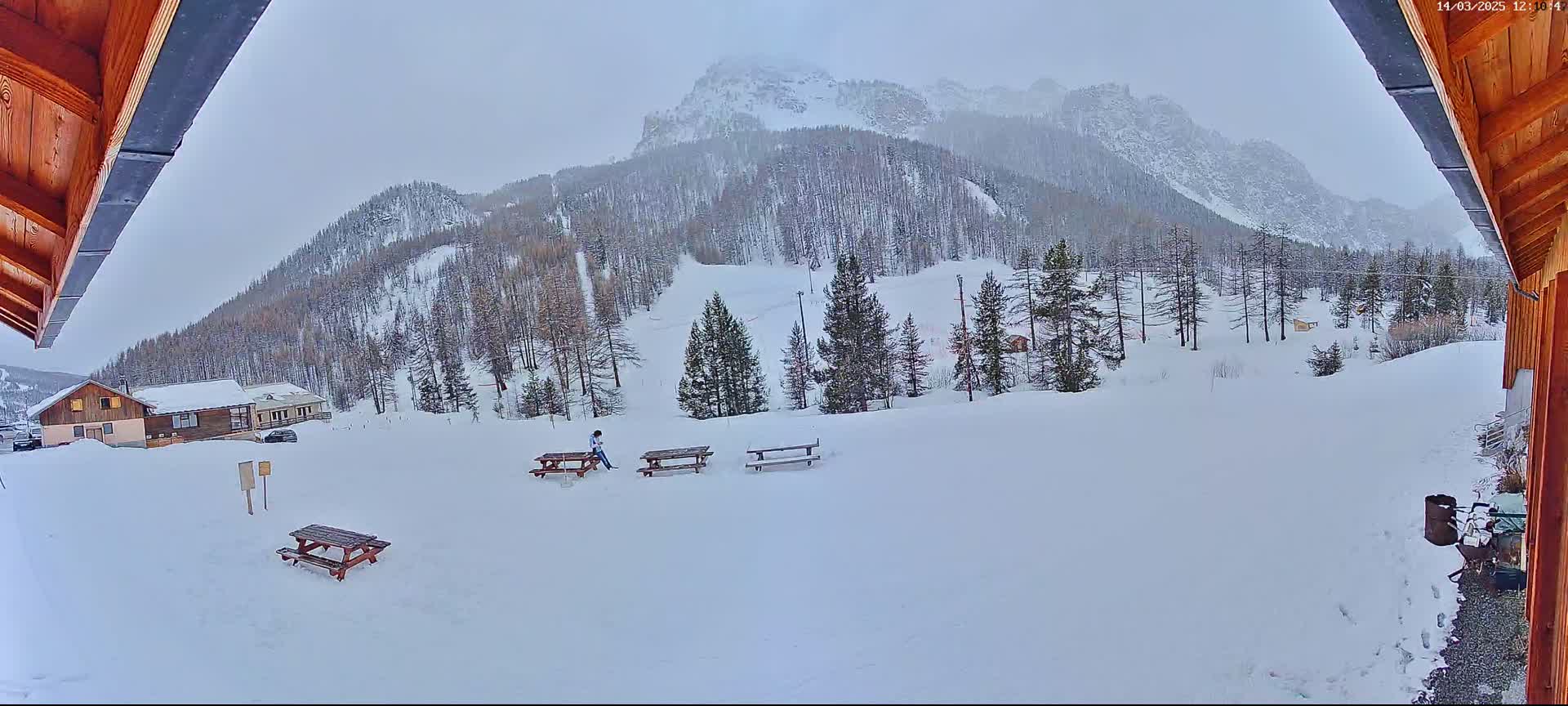 A snowy landscape featuring a snow-covered mountain range, several picnic tables, a few buildings, and a person sitting at one of the tables, viewed from under a wooden overhang.
