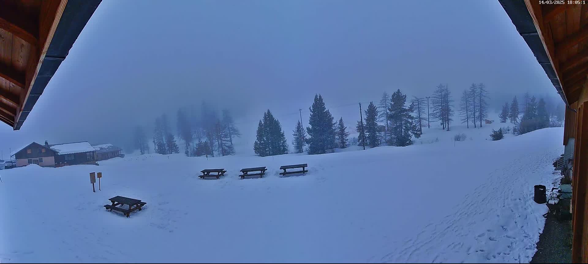 A snowy landscape with several picnic tables, a building in the distance, and a foggy sky is visible from under a wooden overhang.