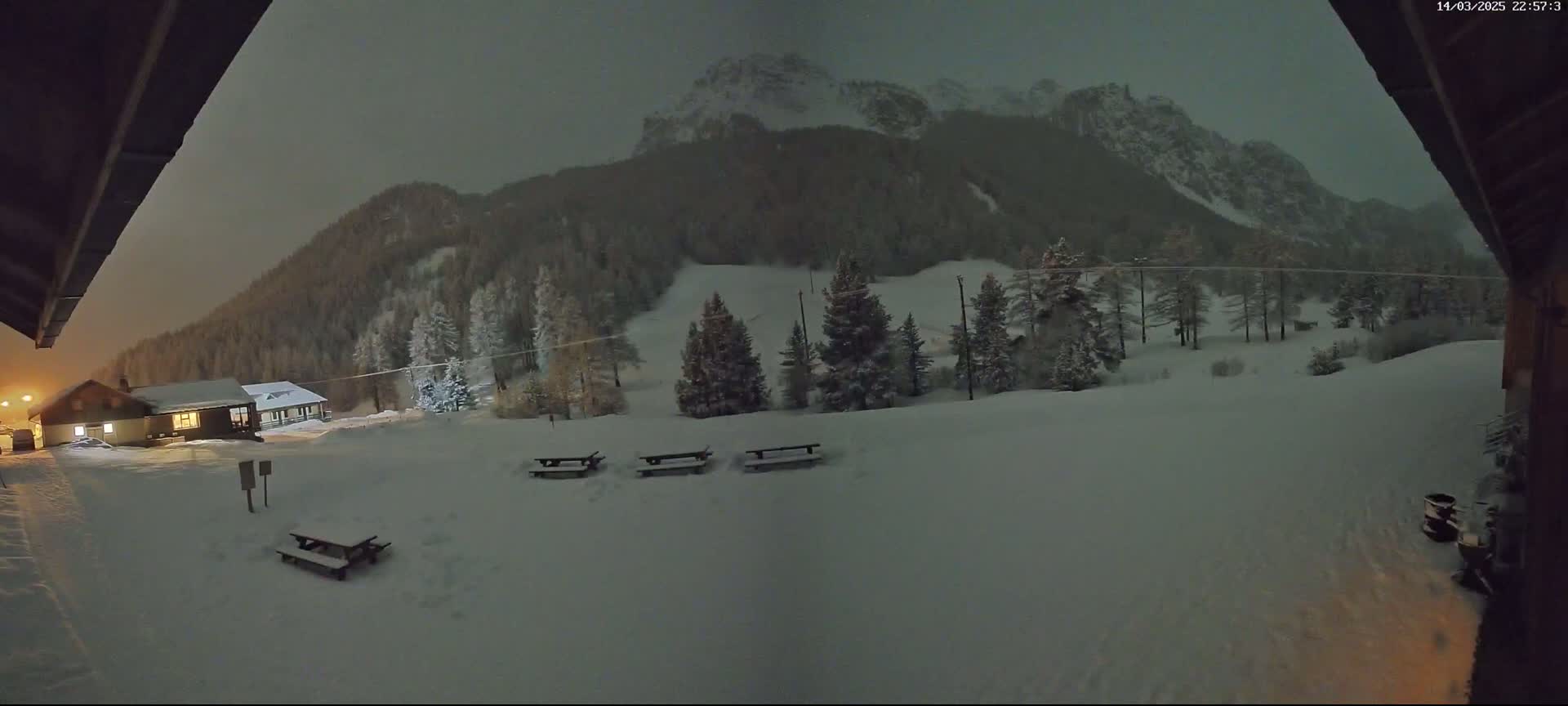 A snow-covered landscape at night, featuring a small building, several picnic tables, and a mountain range in the background under a dark, overcast sky.