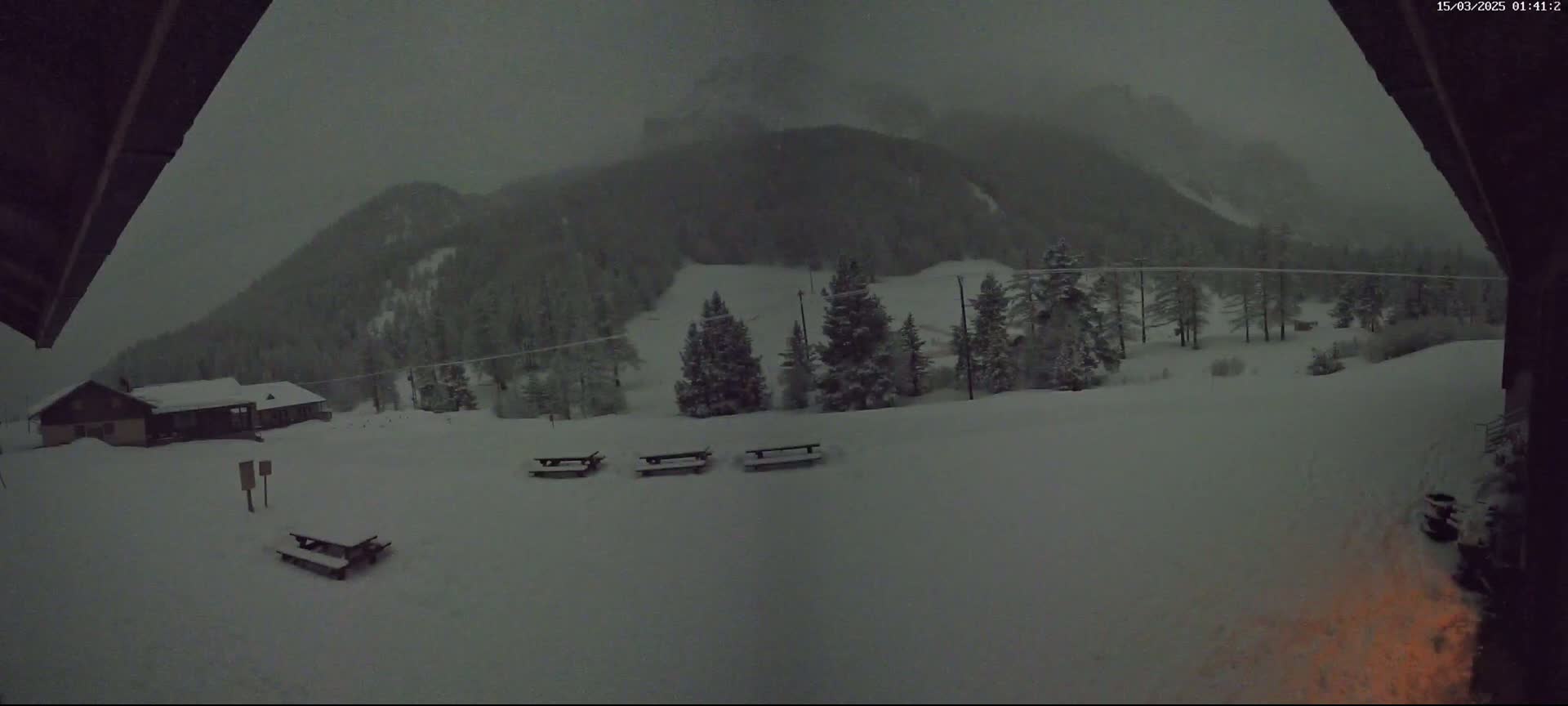 A snow-covered landscape with several picnic tables and a building in the distance is viewed from under a dark overhang during a heavy snowfall.
