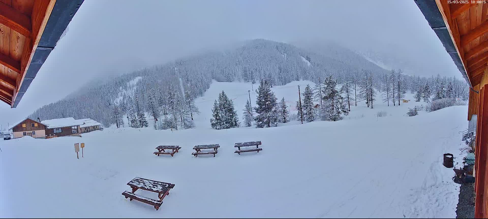A snow-covered landscape with several picnic tables, a building, and a snow-covered mountain in the background under an overcast sky.
