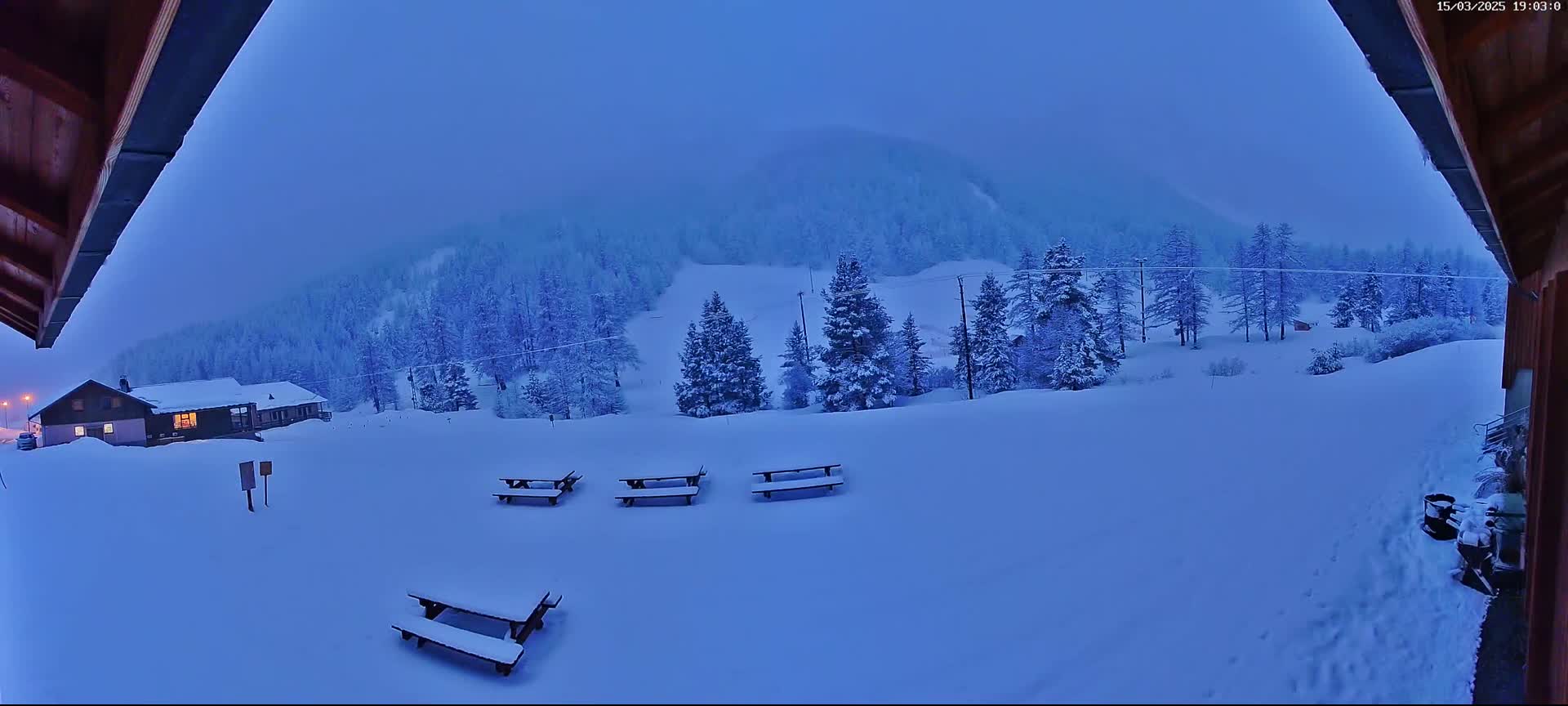 A snow-covered landscape at dusk, featuring several picnic tables, a building with a lit window, and a background of snow-laden trees and a hill.