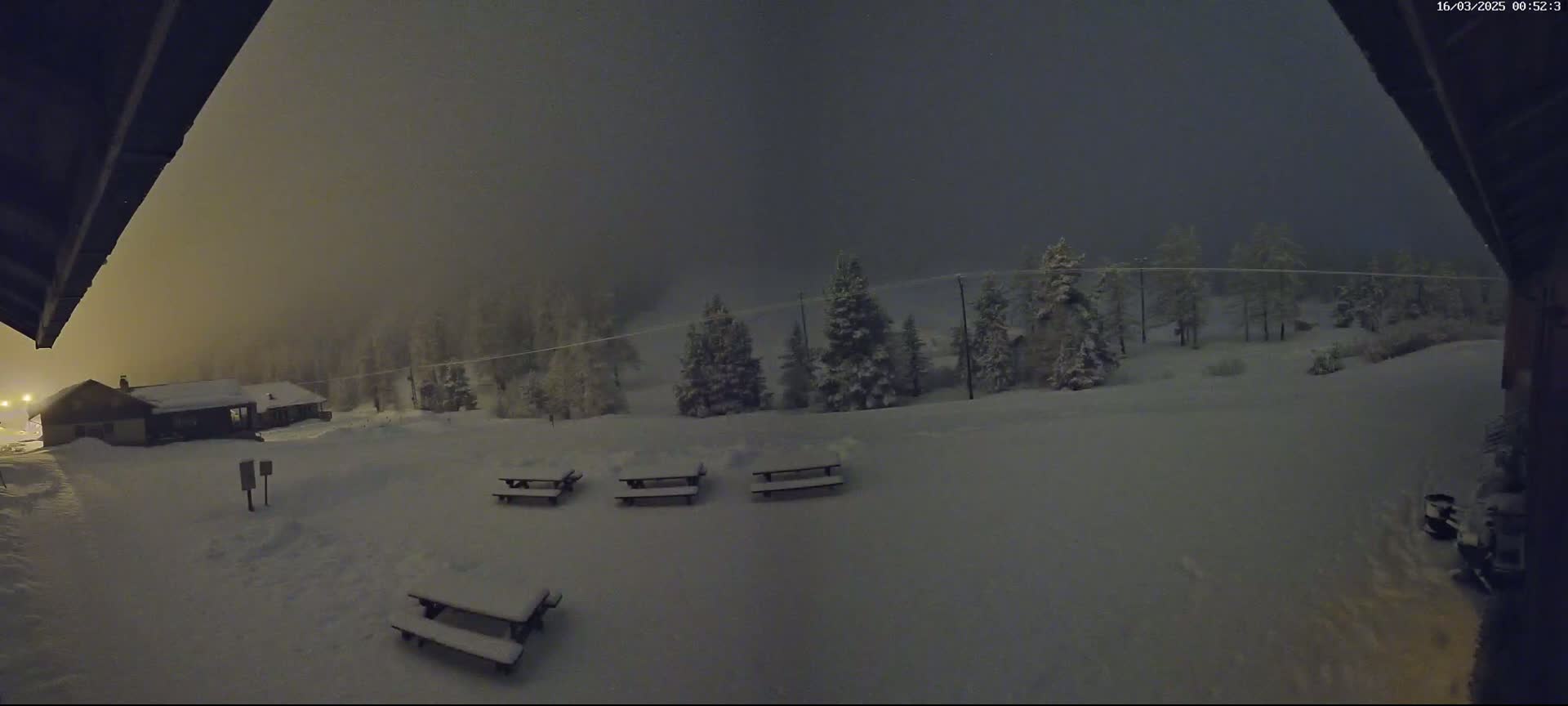 A snow-covered landscape at night, featuring several picnic tables and a building in the distance under a hazy, dark sky.