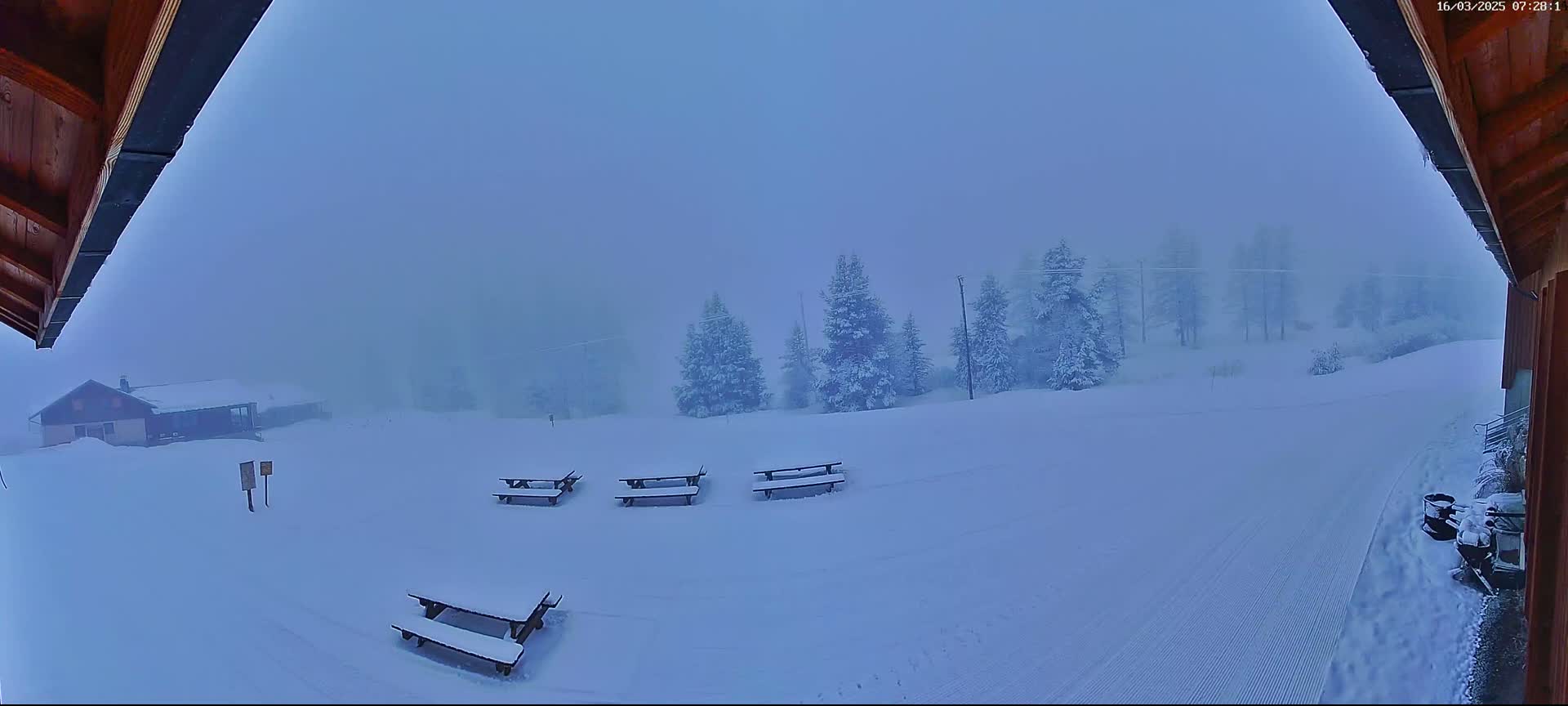 A snow-covered landscape with several picnic tables and a building in the distance is visible through a foggy atmosphere.