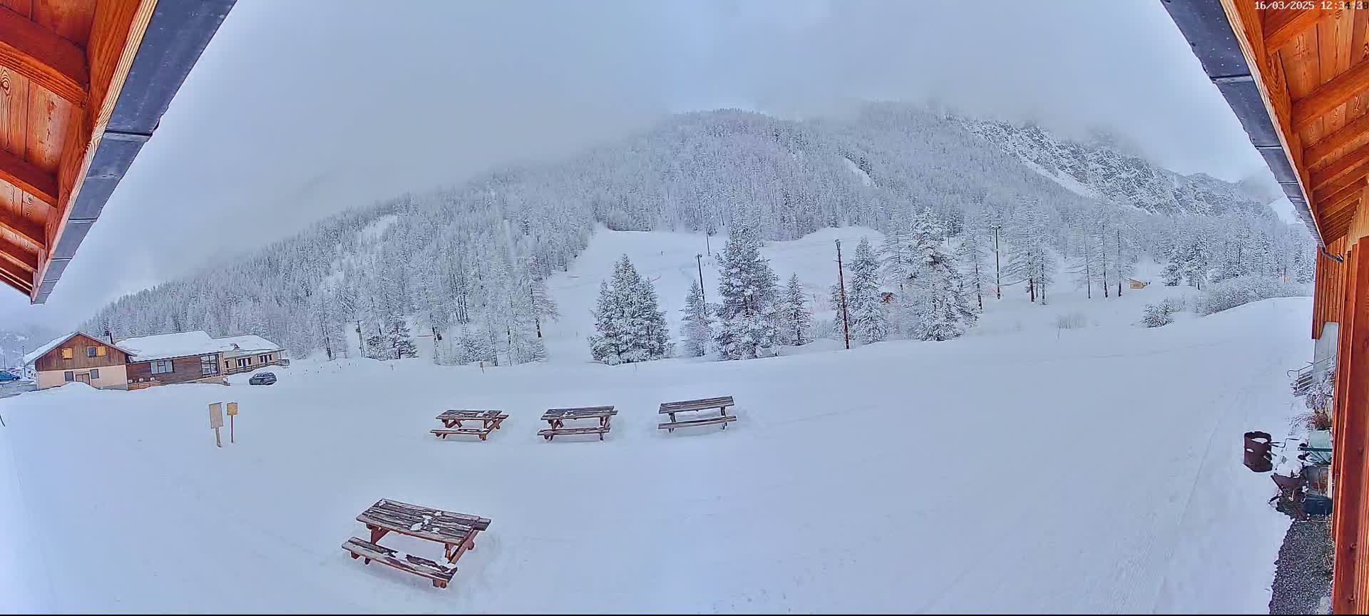 A snow-covered landscape with several picnic tables sits in front of a building, with a snow-covered mountain and forest in the background under an overcast sky.