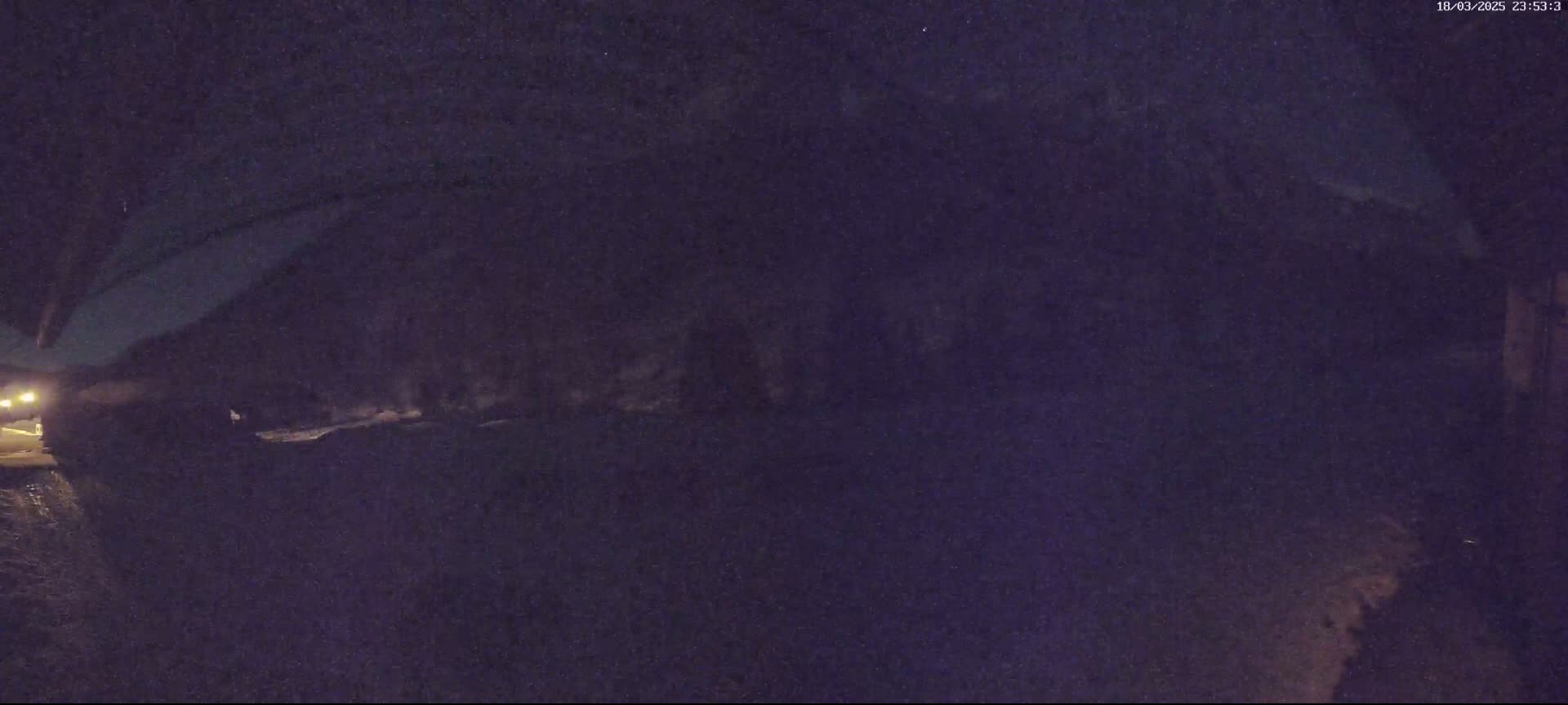 A nighttime, low-light view shows a dark road or path, possibly covered in snow or ice, with a vehicle’s headlights visible in the distance.
