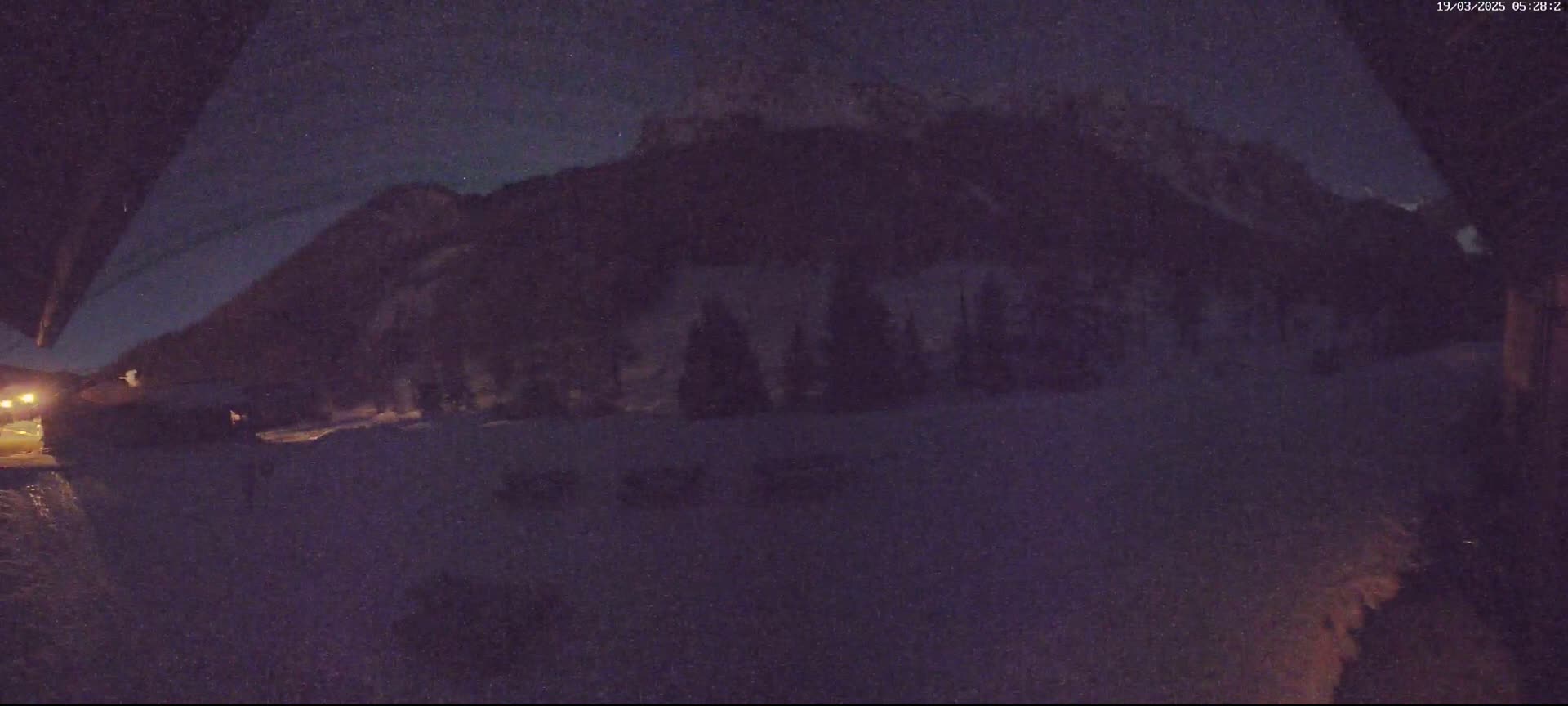 A nighttime, snowy scene shows a dark mountain range under a mostly clear, dark sky, with a few dimly lit structures in the foreground.