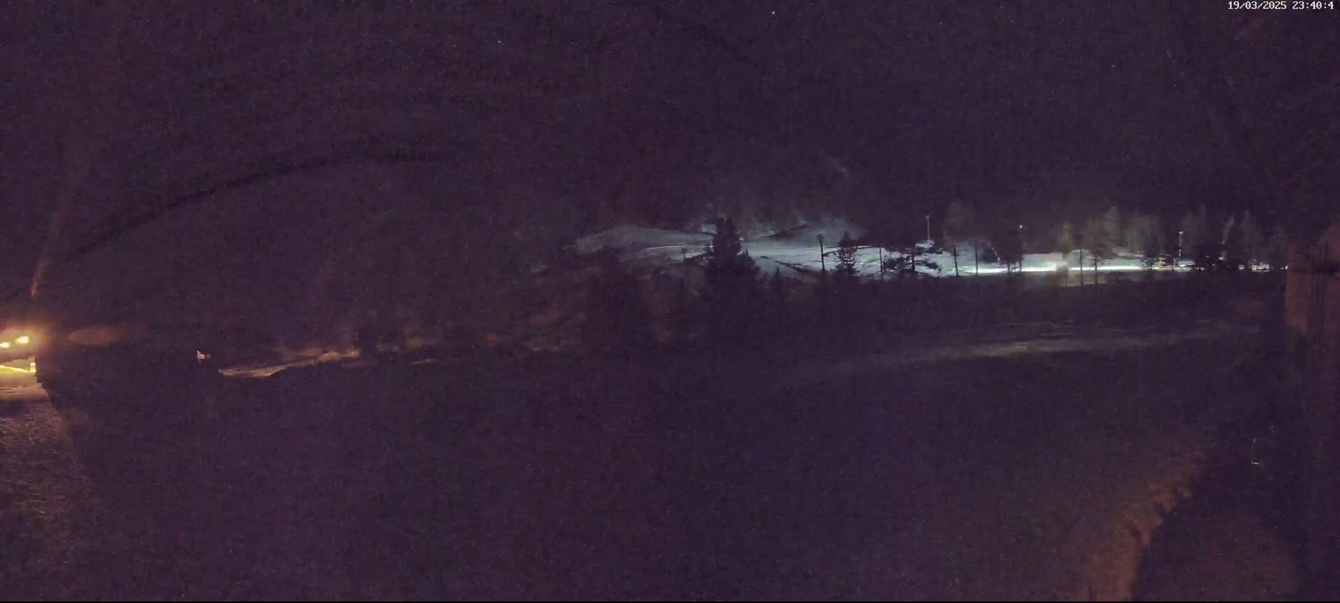 A nighttime, long shot of a snow-covered hillside with sparsely lit trees and a road in the foreground.