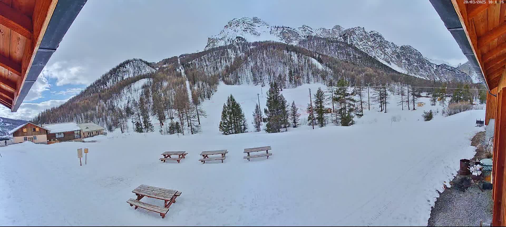 A snowy mountainside view with several picnic tables in the foreground, seen from under a wooden overhang.