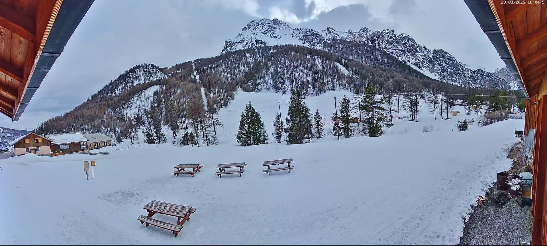 A snowy mountain landscape is viewed from a partially covered outdoor patio with several picnic tables, under an overcast sky.