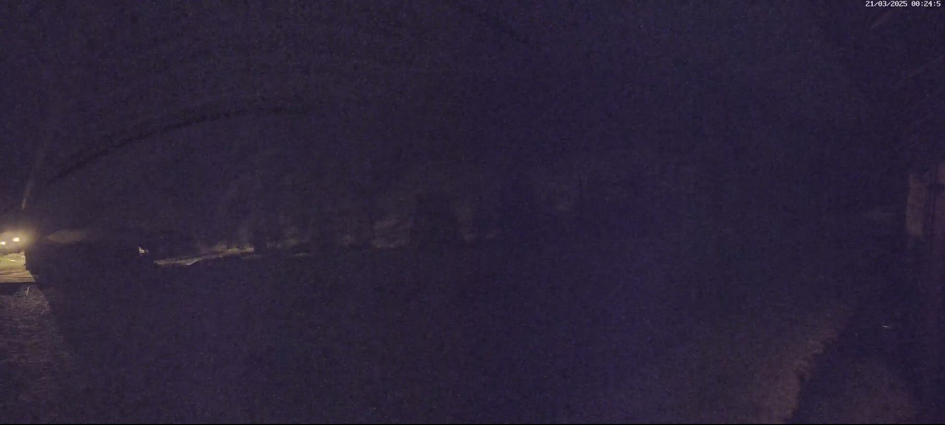 A low-light nighttime image shows a dark, shadowy area under what appears to be an overpass or bridge, with a faint light visible in the distance.