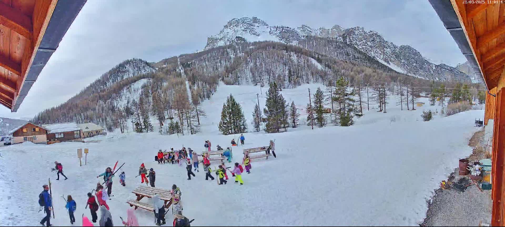 A snowy mountainside scene with several people gathered near picnic tables, some carrying skis, under an overcast sky.