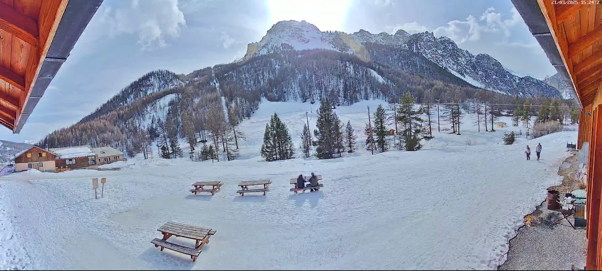 A sunny, snow-covered mountain landscape is viewed from a wooden structure, with several picnic tables and a few people visible in the foreground.