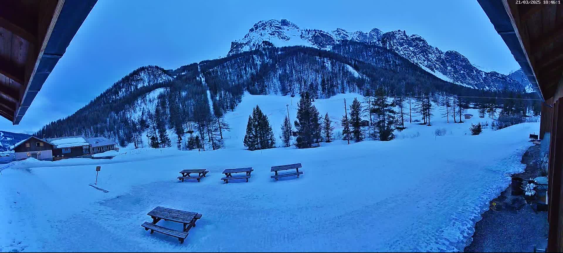 Four picnic tables sit in a snow-covered area overlooking a snow-dusted mountain range under a twilight sky.