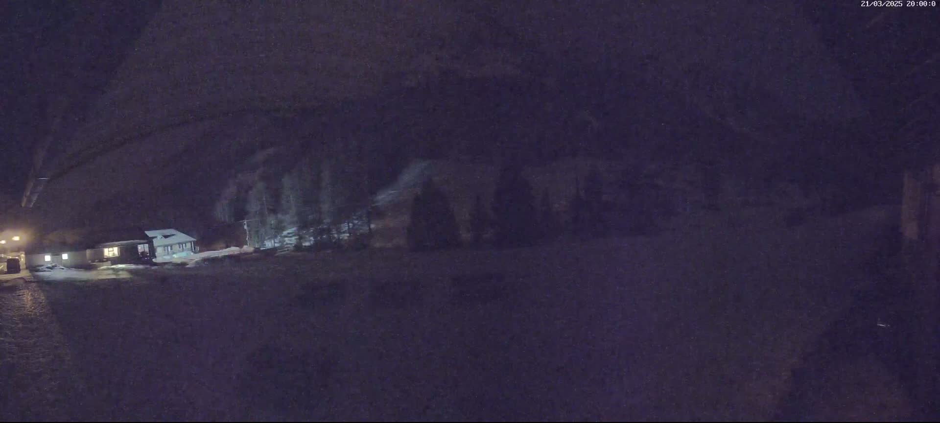 A nighttime, snowy scene shows a house and trees under a dark sky.