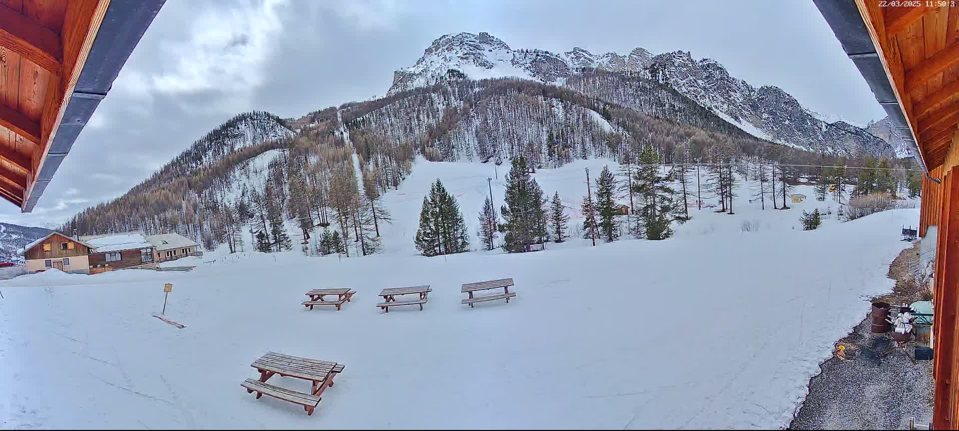 A snow-covered landscape with several picnic tables in the foreground and a mountain range in the background under a cloudy sky.
