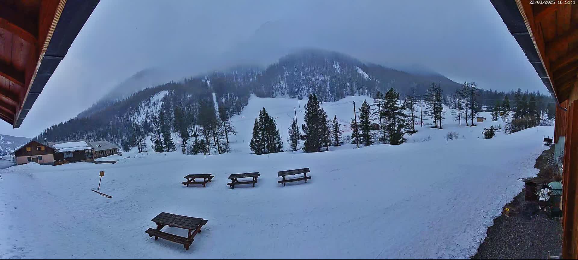 A snowy landscape with several picnic tables is visible from a wooden structure's edge, overlooking a snow-covered hillside and distant evergreen trees under an overcast sky.