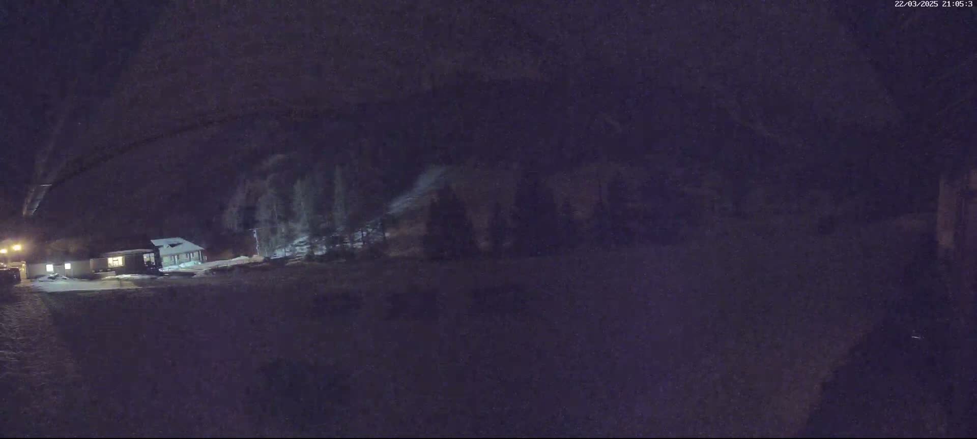A snow-covered landscape at night shows a small building illuminated with lights, and trees behind it under a dark sky.