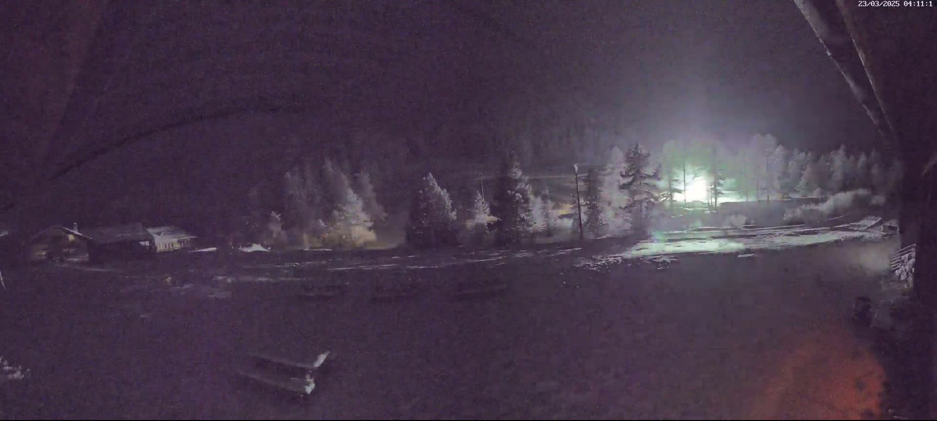 A nighttime, snowy scene shows a few buildings and trees illuminated by a bright light in the distance.