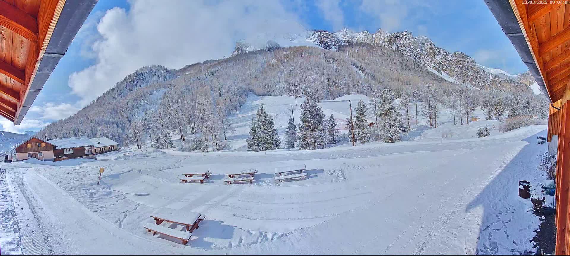 A snow-covered landscape with several picnic tables, a small building, and a mountain in the background under a partly cloudy sky.
