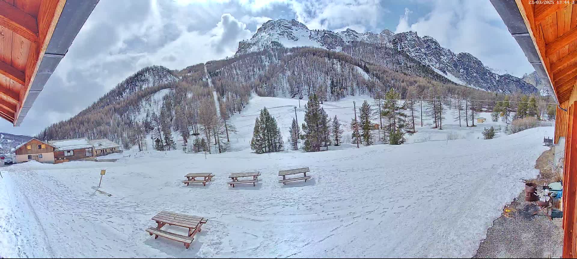 A snow-covered mountain landscape with several picnic tables and a building is visible from under a wooden overhang on a partly cloudy day.