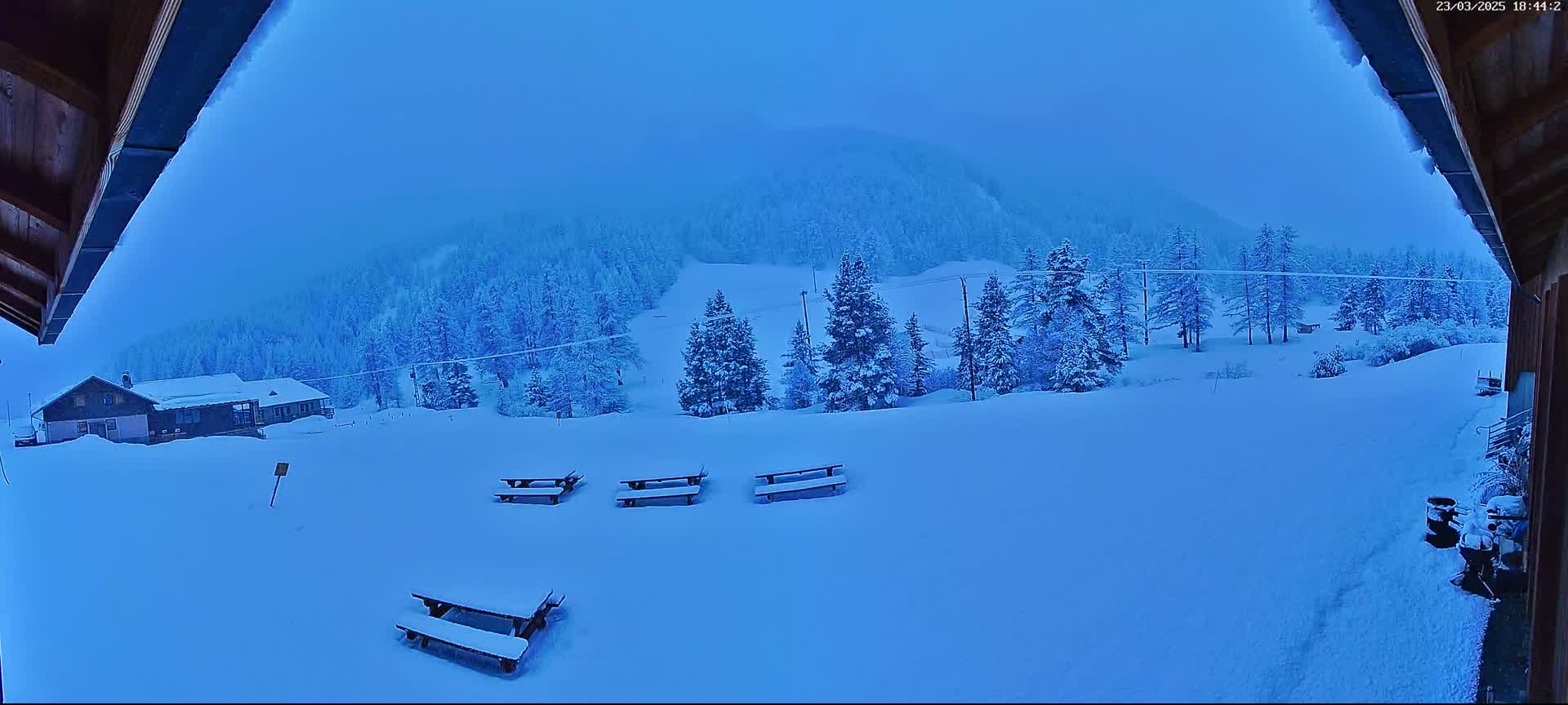 A snow-covered landscape with several picnic tables and a building is visible from a viewpoint under an overhanging roof in overcast conditions.