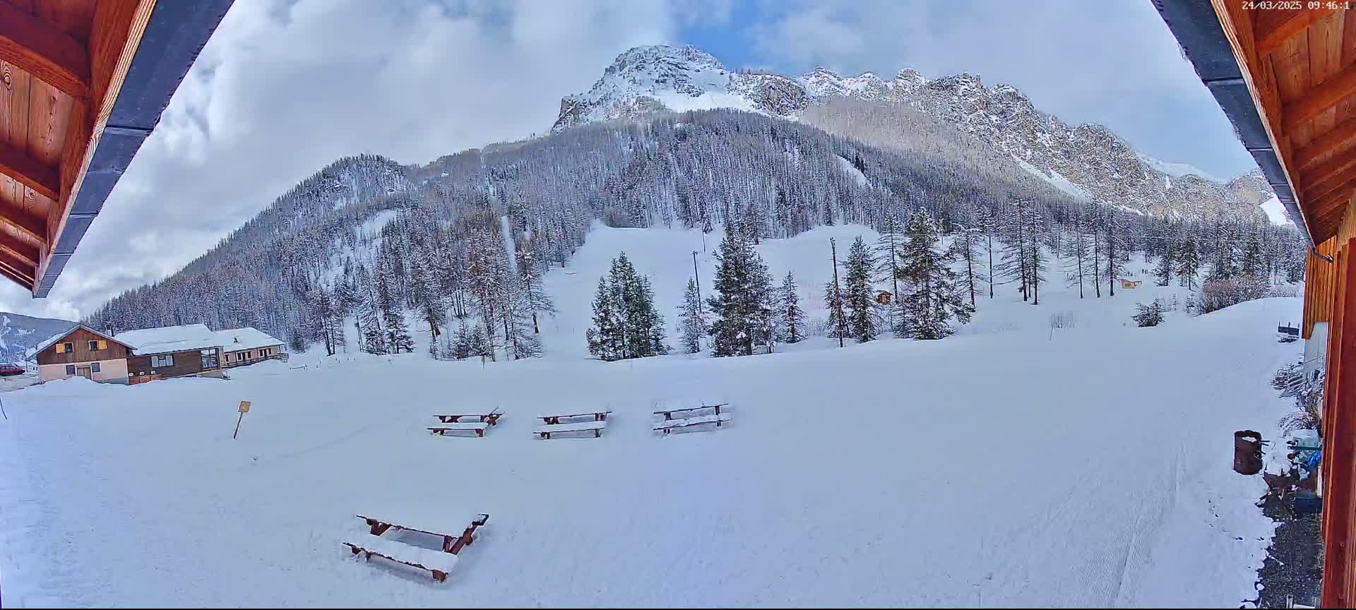 A snow-covered landscape with several picnic tables, a building, and a snow-capped mountain in the background under a partly cloudy sky.