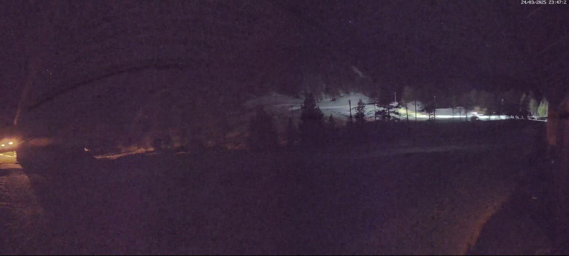 A nighttime view of a snow-covered area, sparsely lit, with trees in the background and a dark foreground.
