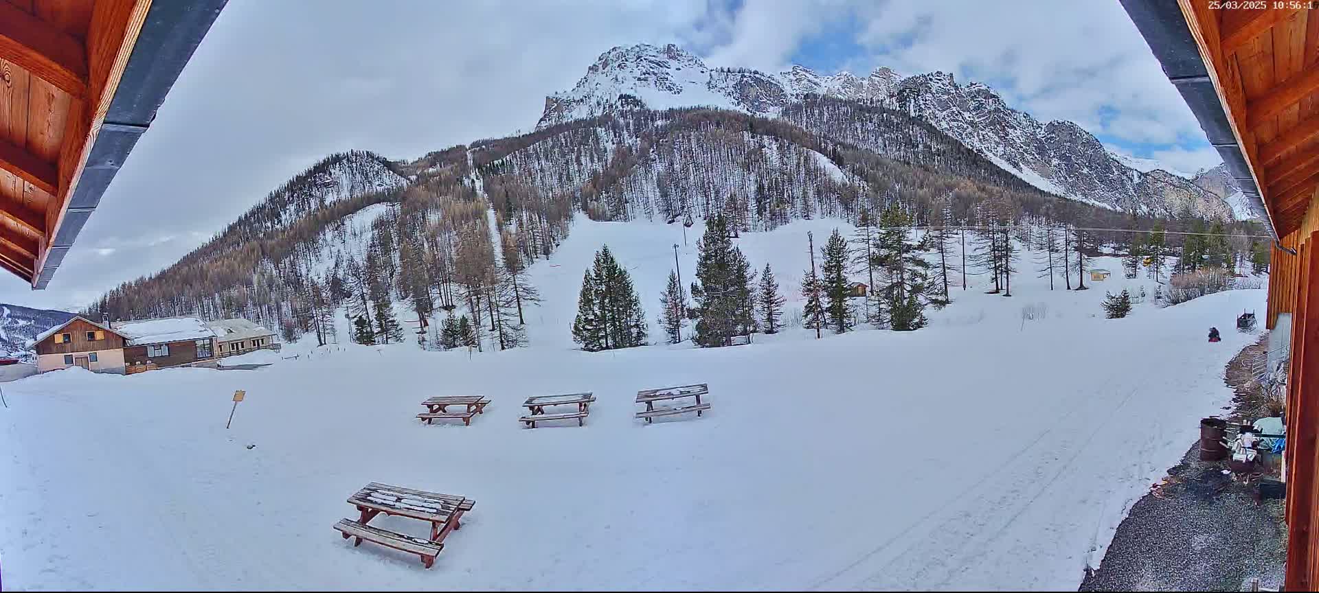 A snow-covered mountainside with several picnic tables and a small building is visible from a partially enclosed wooden structure on an overcast day.
