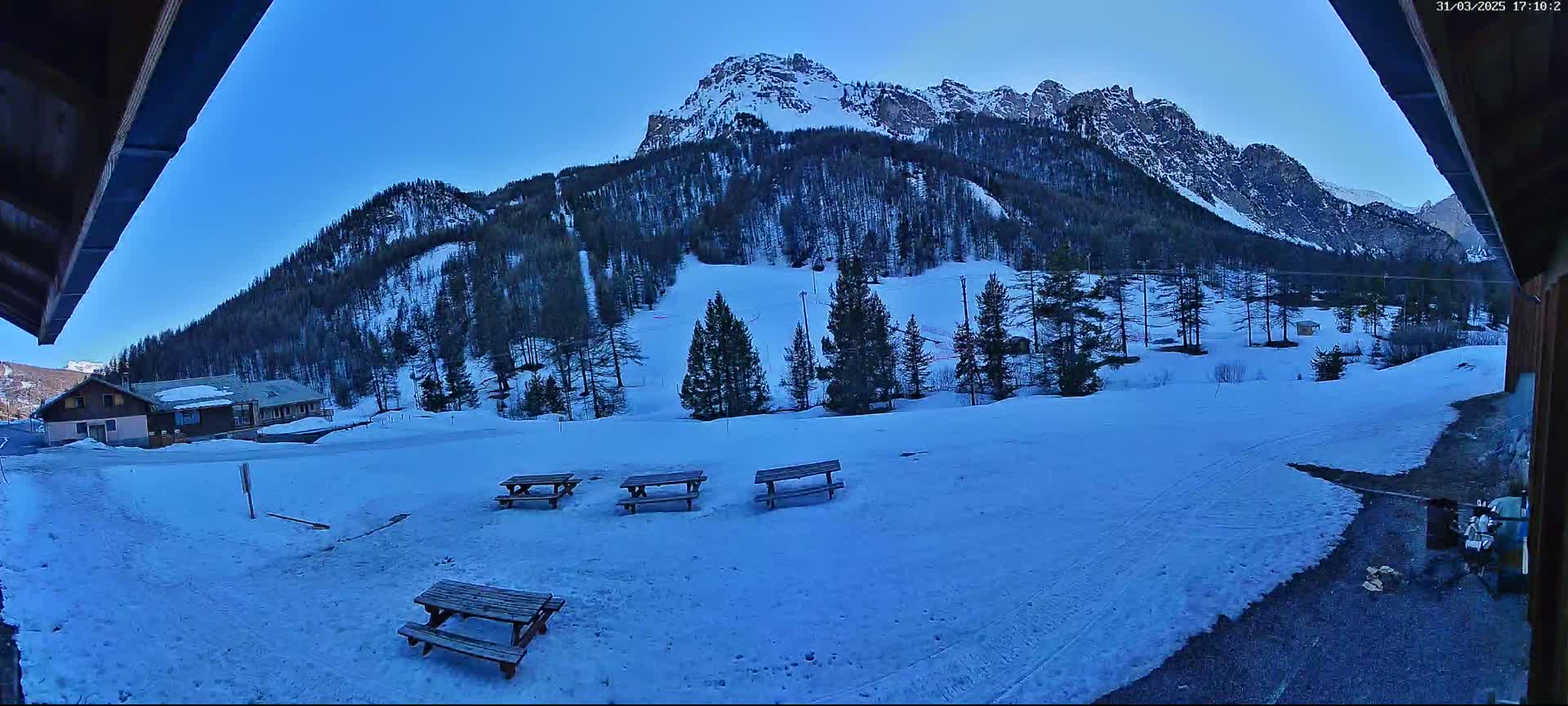 A snow-covered landscape with several picnic tables is viewed from under a wooden overhang, featuring a mountain range in the background under a clear blue sky.