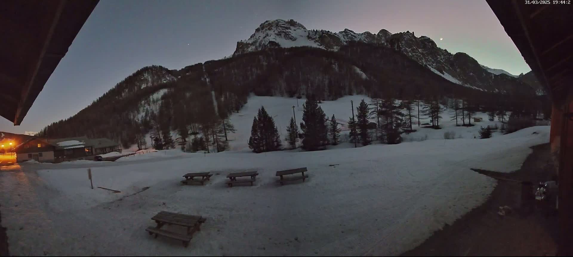 Four picnic tables sit in a snow-covered area at twilight, with a snow-capped mountain and dark pine trees in the background.