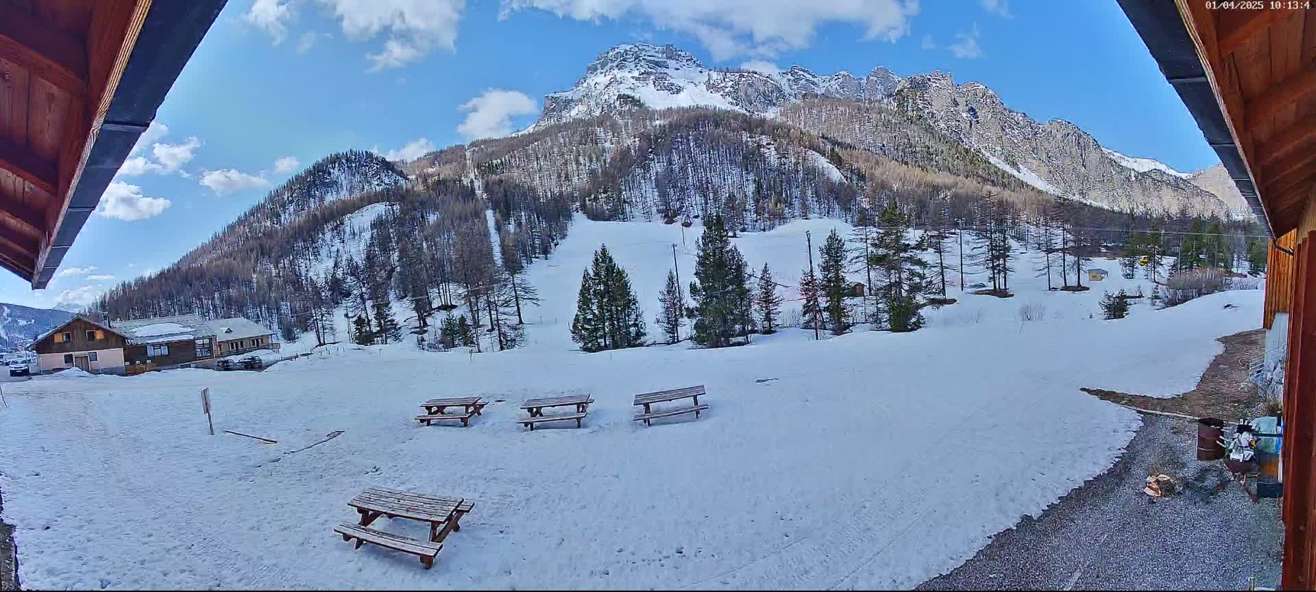 A snow-covered mountainside with several picnic tables sits in front of a small building, under a partly cloudy sky.