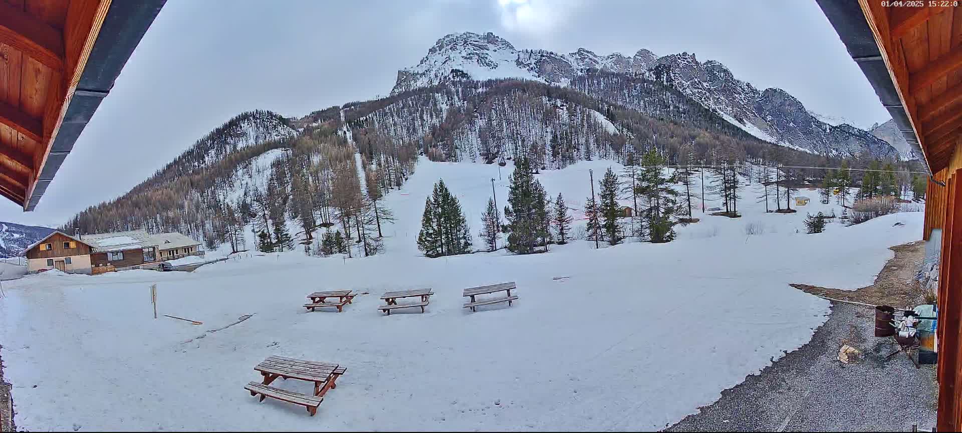 A snow-covered landscape with several picnic tables, a building, and a mountain range in the background under a cloudy sky.