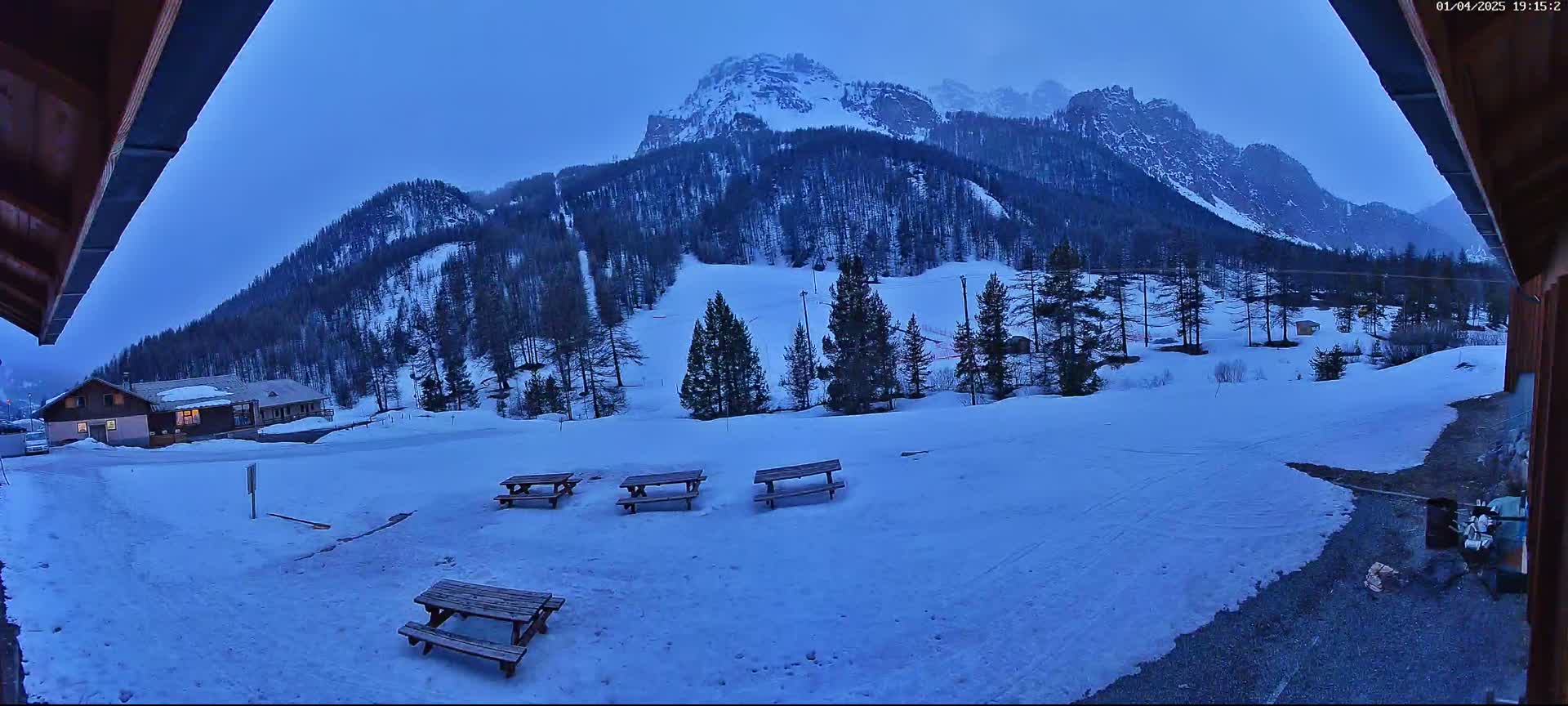 Four picnic tables sit in a snow-covered area overlooking a snow-covered mountain range under a twilight sky.