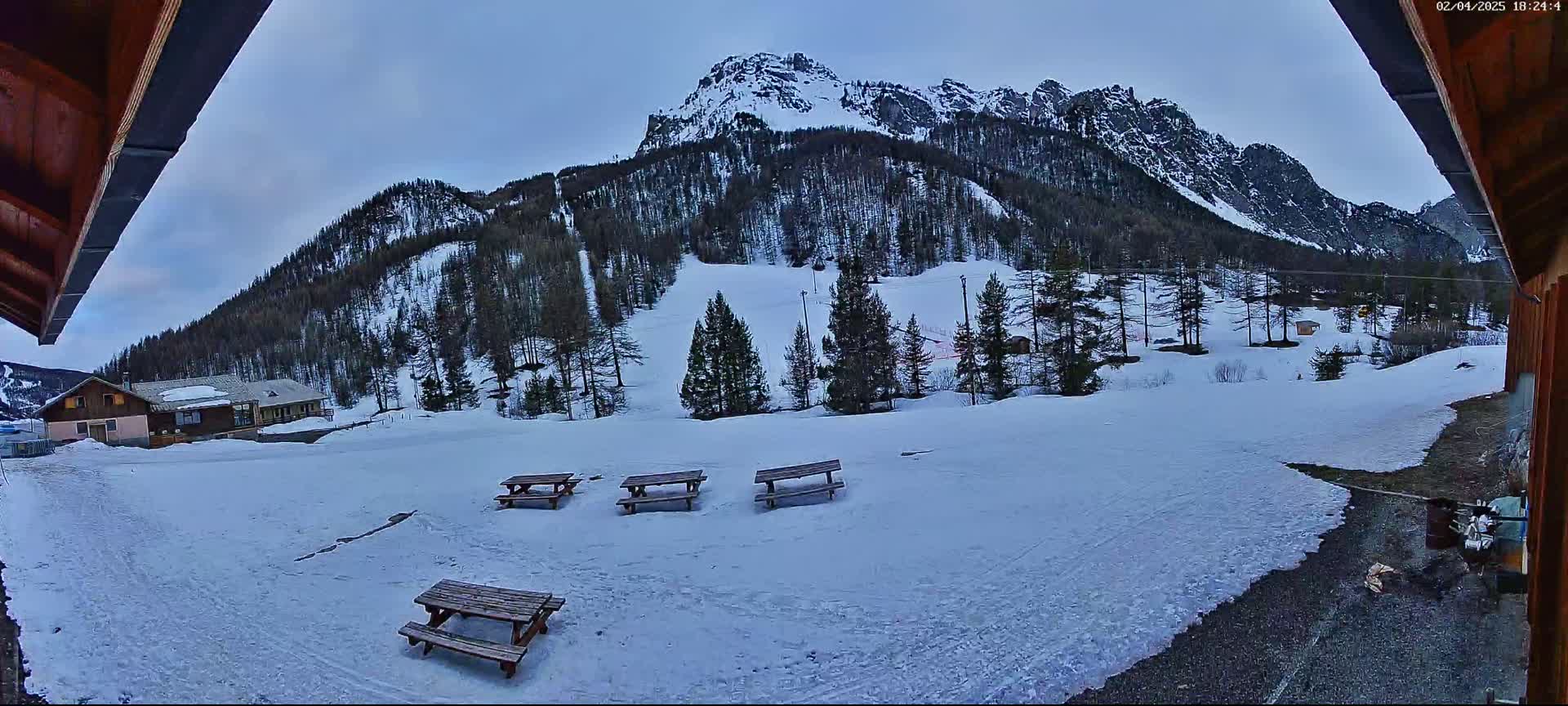 A snow-covered landscape with several picnic tables in the foreground, a building in the mid-ground, and a snow-capped mountain range under a twilight sky.