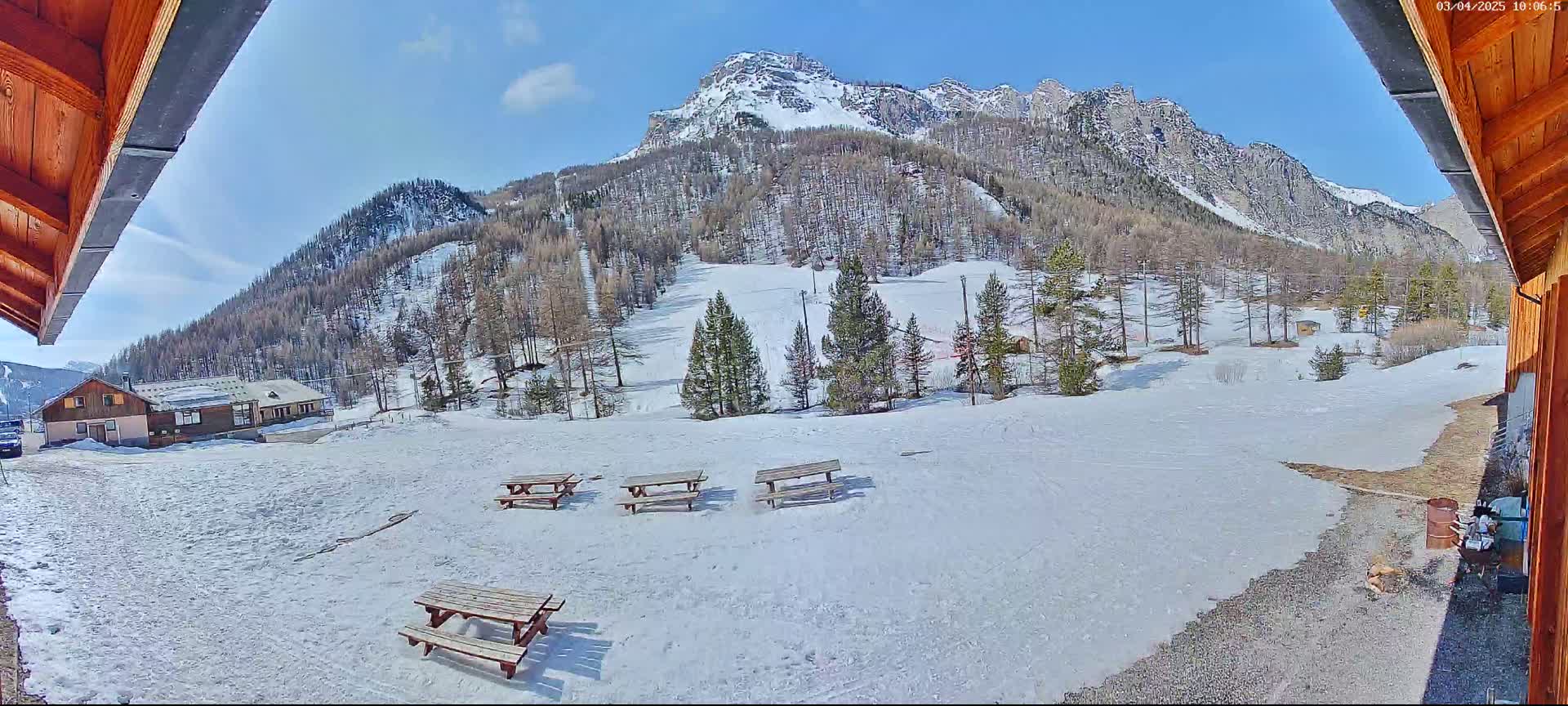 A sunny view from under a wooden overhang shows a snow-covered area with several picnic tables, a small building in the distance, and a mountain range in the background.