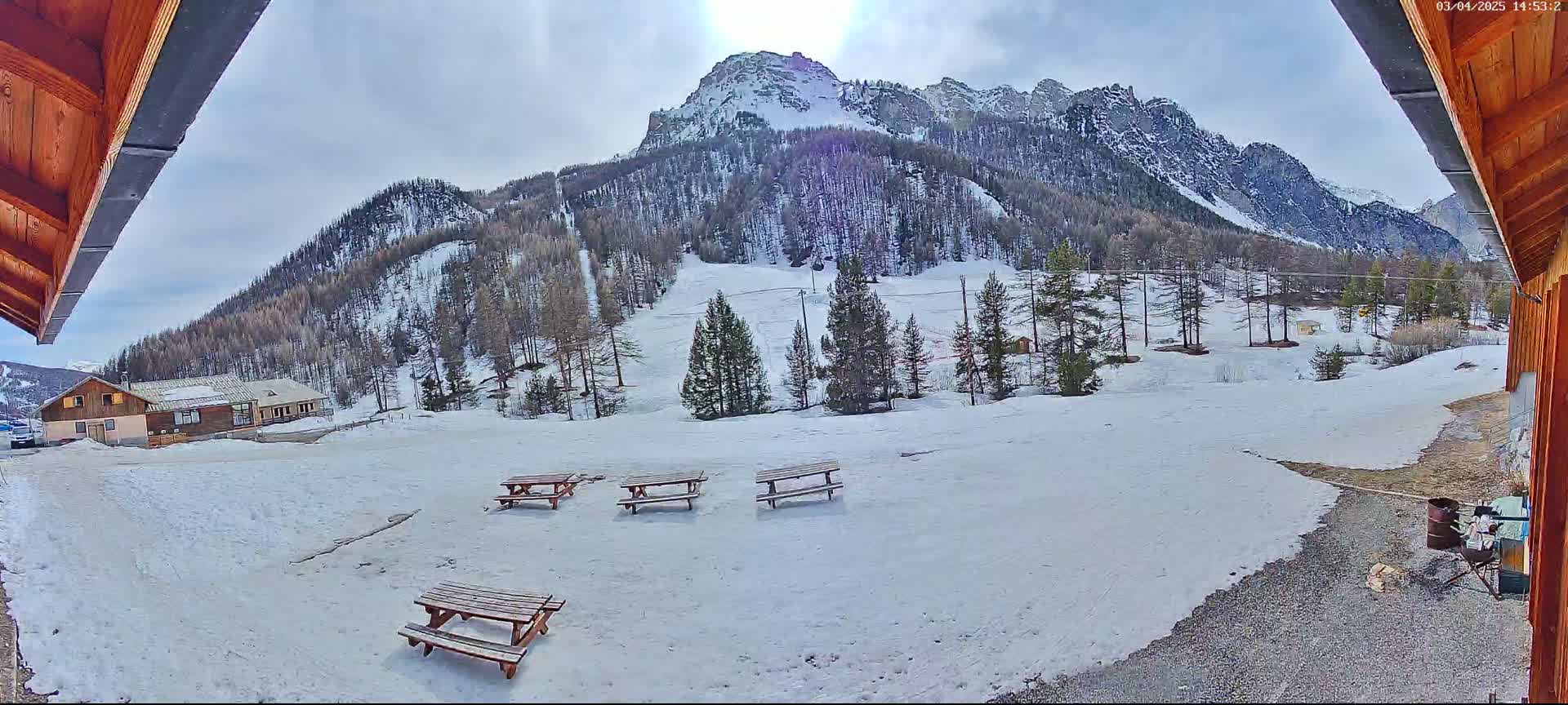 A snowy mountain landscape is viewed from a wooden structure's exterior, showing several picnic tables in the foreground and a building in the midground, under a mostly cloudy sky.